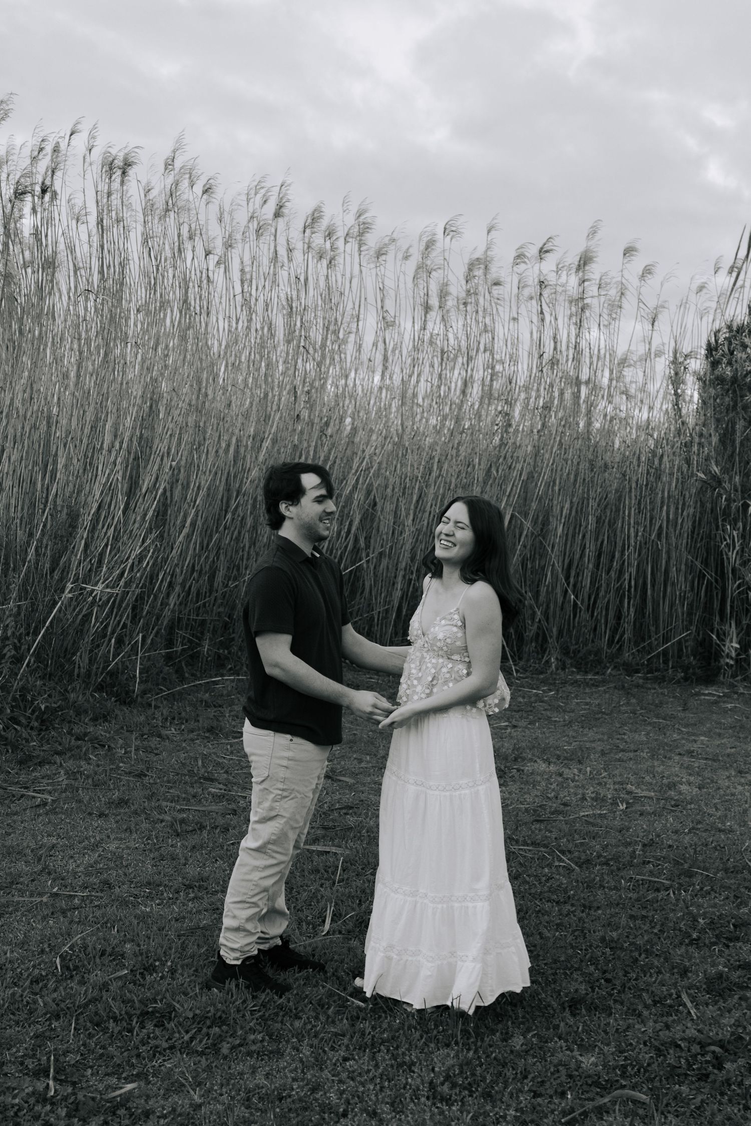 Black and white photo of couple holding hands and laughing together in front of tall wild reeds.