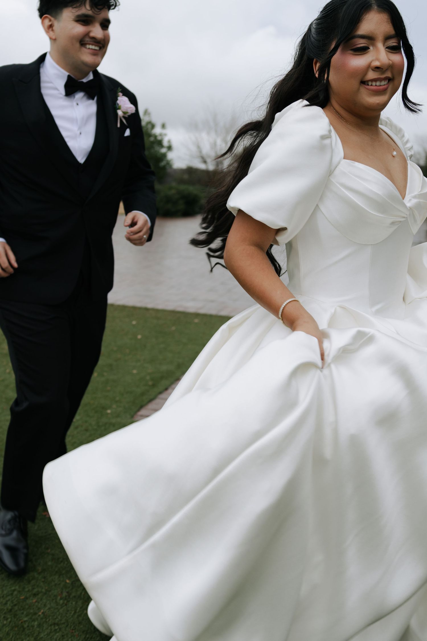 Smiling bride in white structured gown runs joyfully alongside partner in black tuxedo on a cloudy outdoor setting.