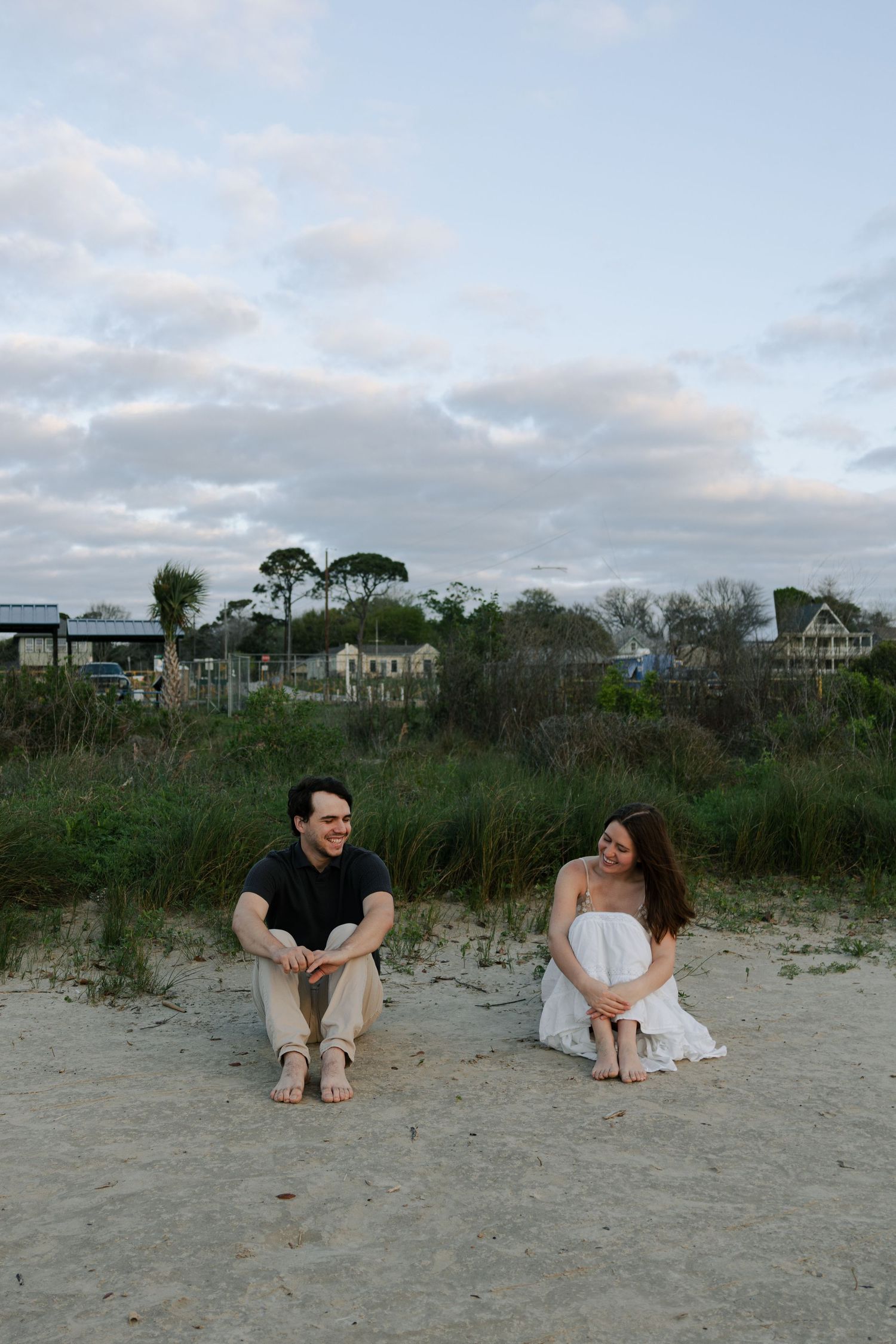 Engaged couple sitting apart on sandy beach smiling at each other with coastal homes visible in background.