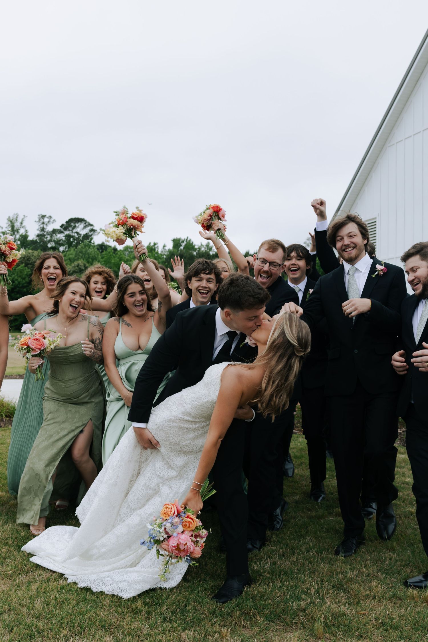 Wedding party cheering as bride and groom share a romantic dip kiss outdoors near a white barn.