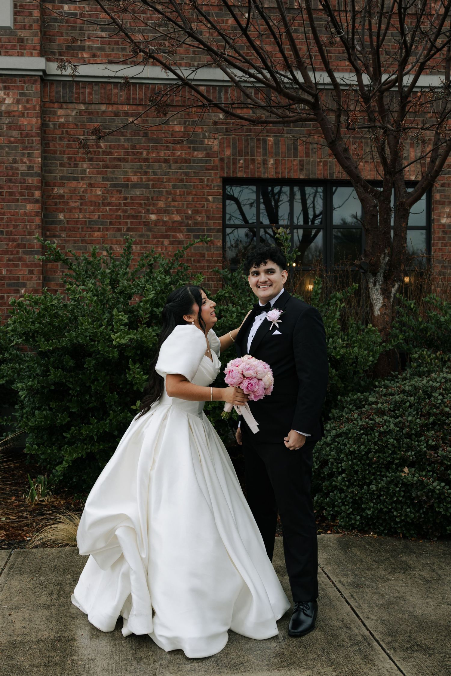 Bride in white gown and groom in black tuxedo pose together outdoors holding pink bouquet before brick building.