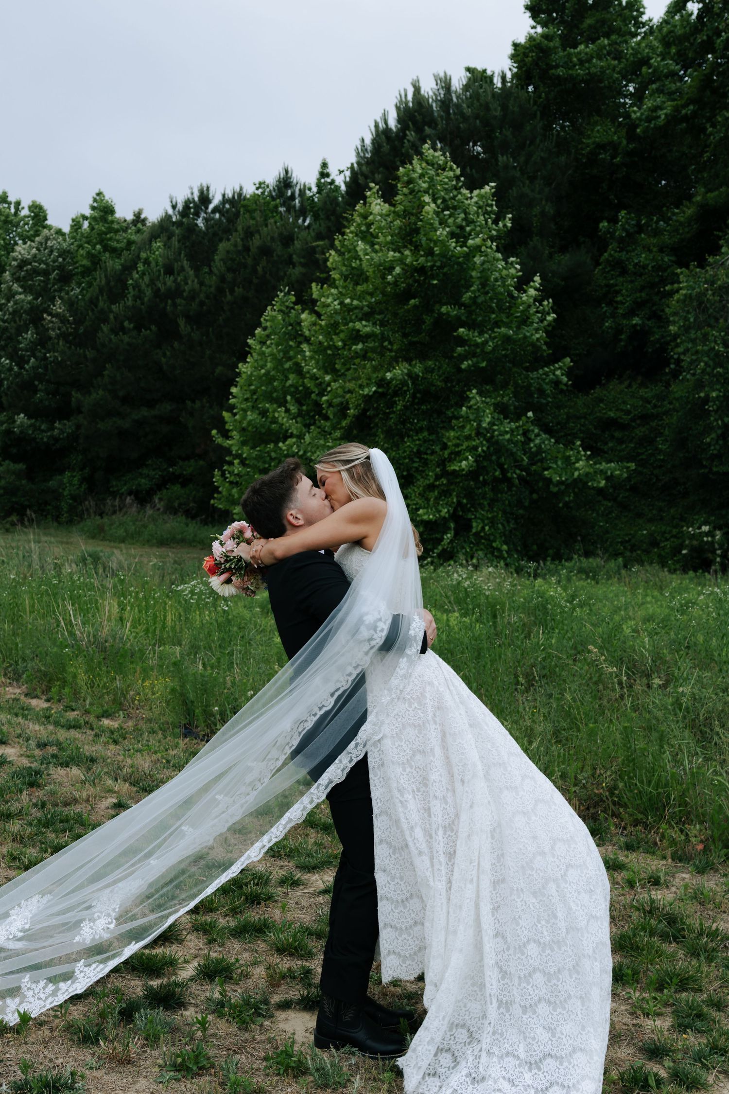 Groom lifts bride in lace gown and veil for romantic kiss in green field.