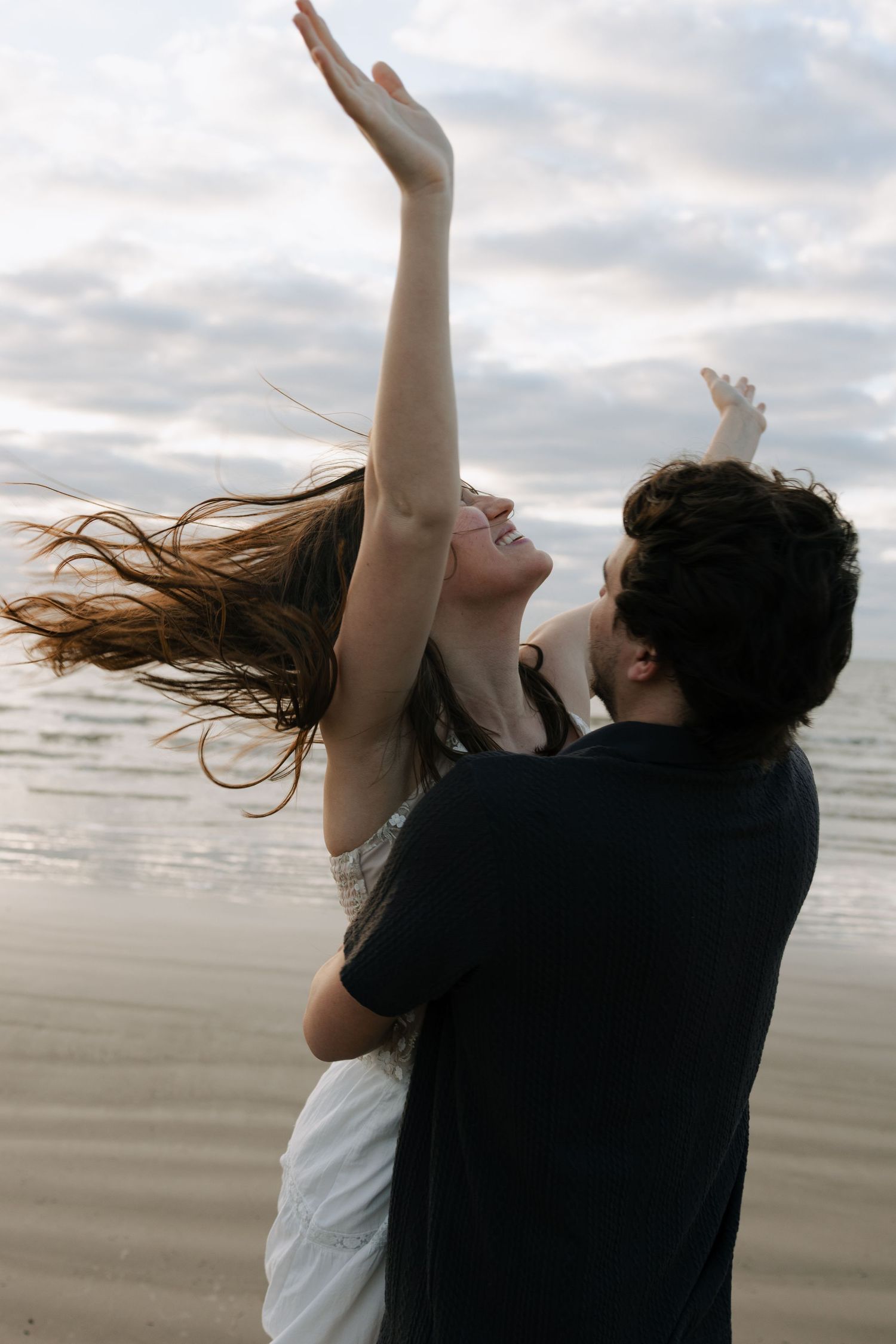 Couple dancing joyfully on a beach at sunset, woman with flowing hair reaching arms up toward cloudy sky.