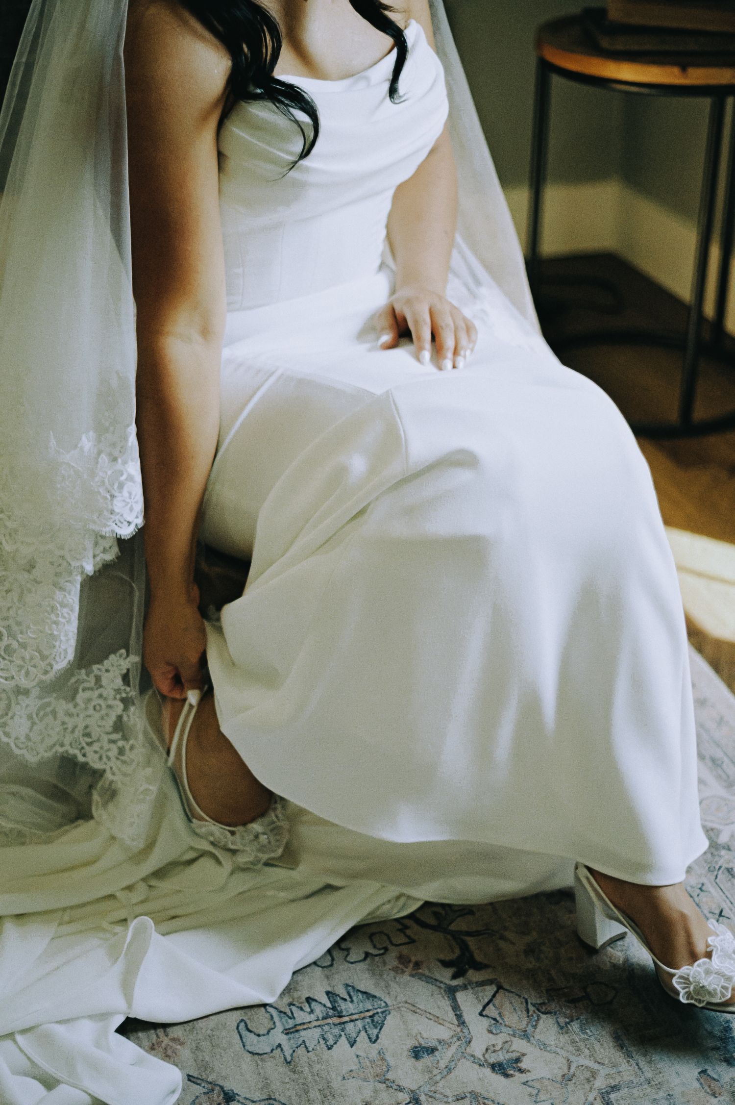 Bride in white satin wedding gown and veil adjusting her heel shoe while seated, showcasing elegant bridal style.