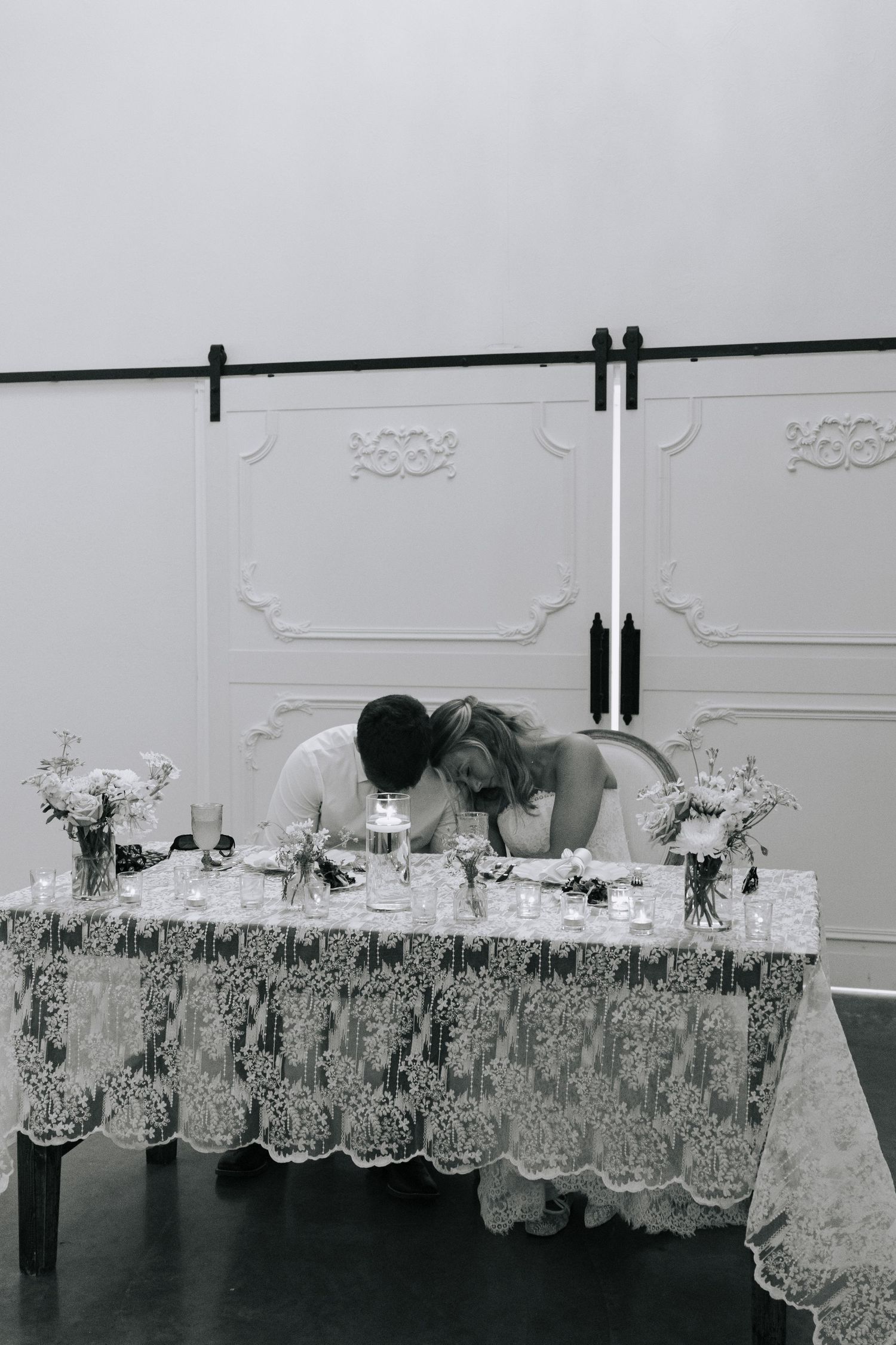 Black and white photo of bride and groom sharing an intimate moment at their elegant lace-covered sweetheart table.