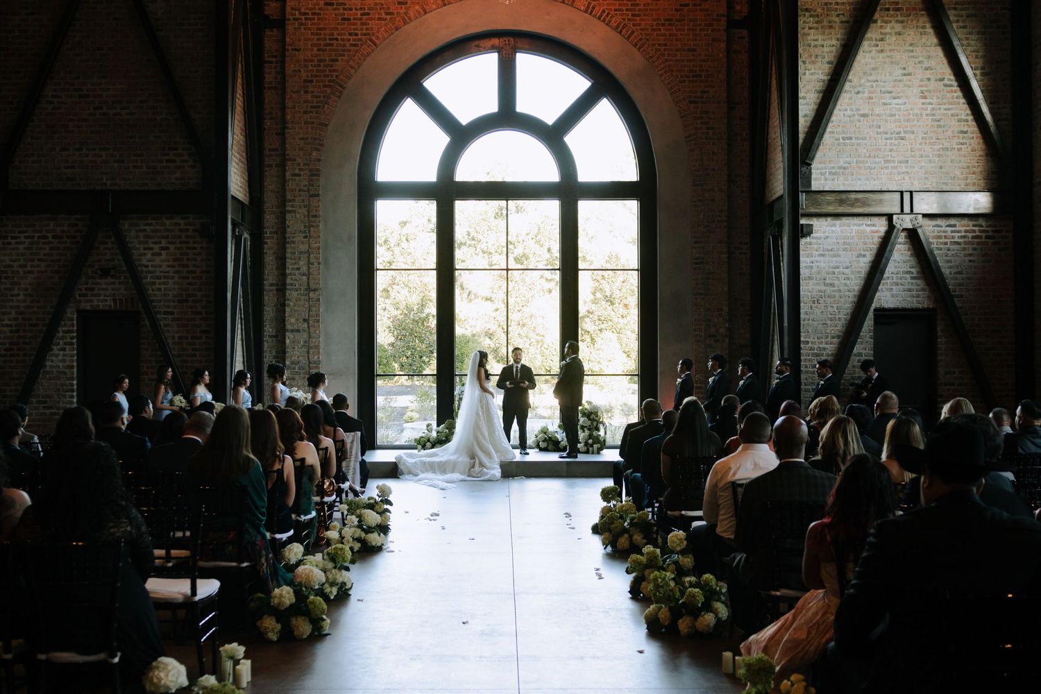Elegant wedding ceremony in industrial venue with arched window, floral aisle, and guests seated on both sides.