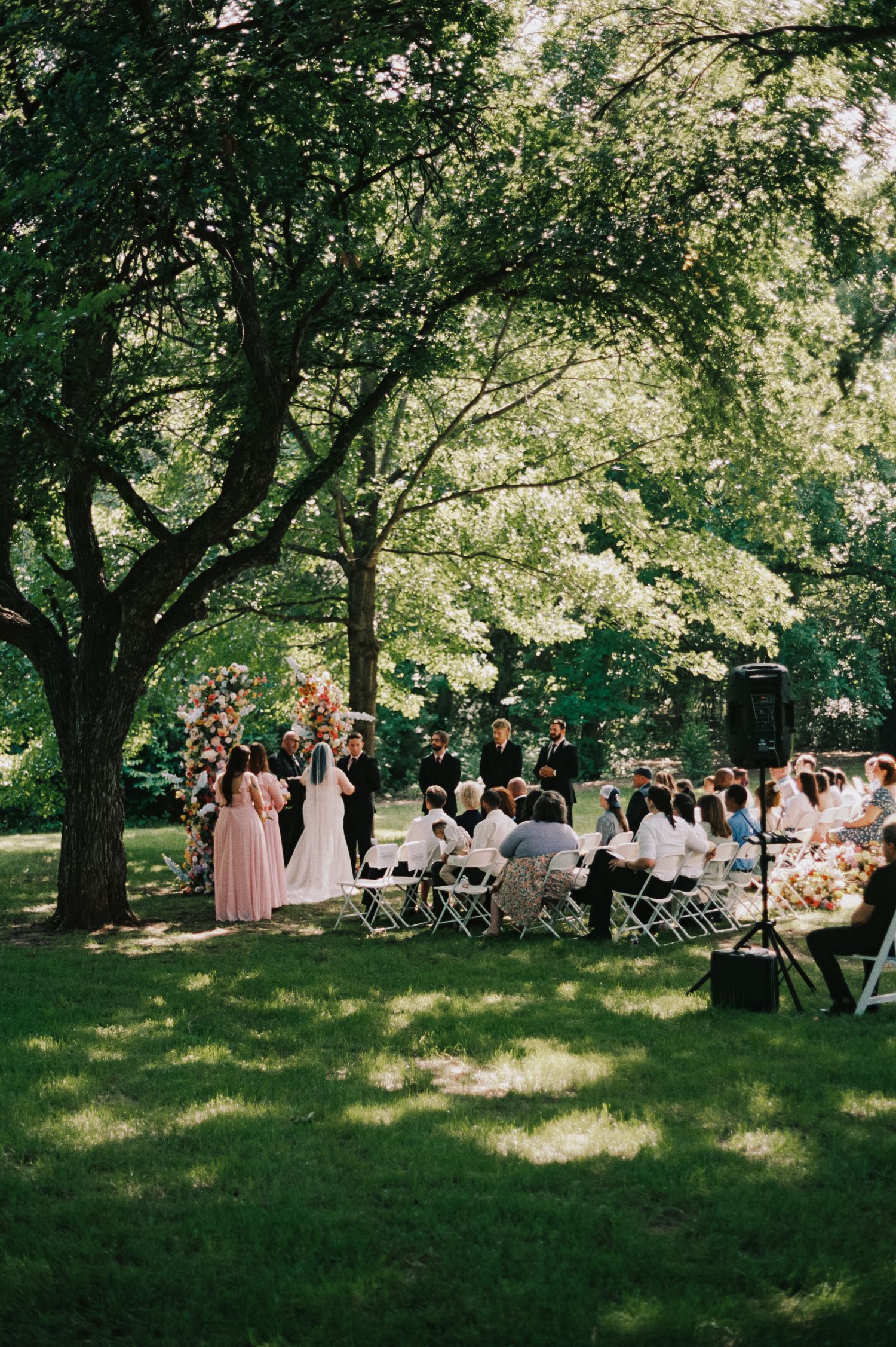 Outdoor garden wedding ceremony under lush green trees with guests seated on white chairs facing the bride and groom.