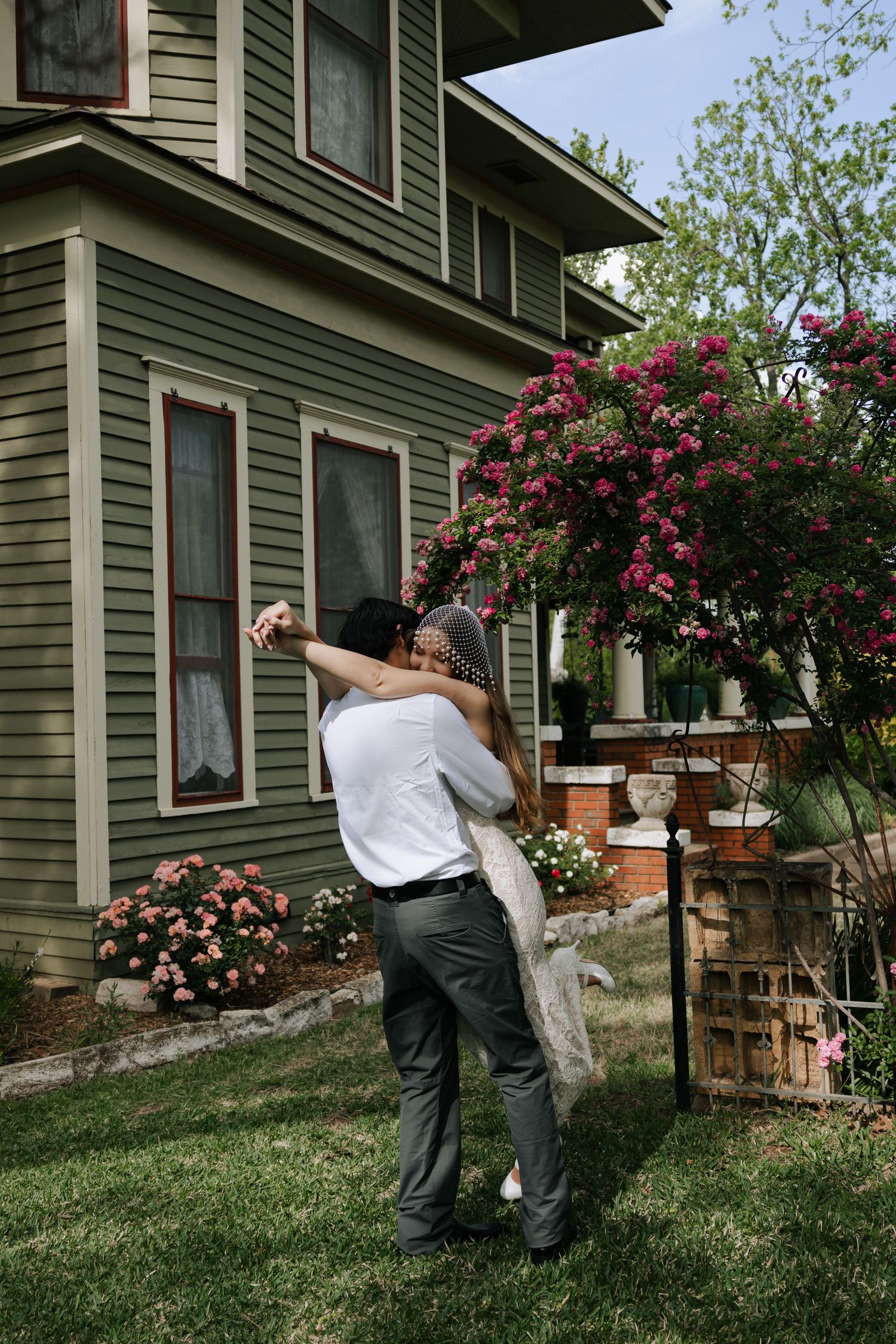Groom lifts bride in a joyful embrace outside a charming green craftsman house surrounded by blooming pink roses.