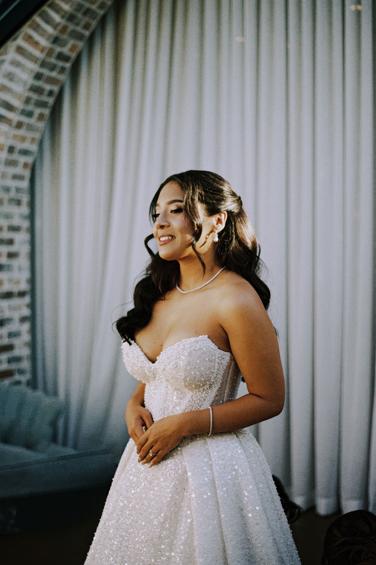 Radiant bride in strapless beaded ball gown with pearl jewelry smiling in softly lit wedding venue.