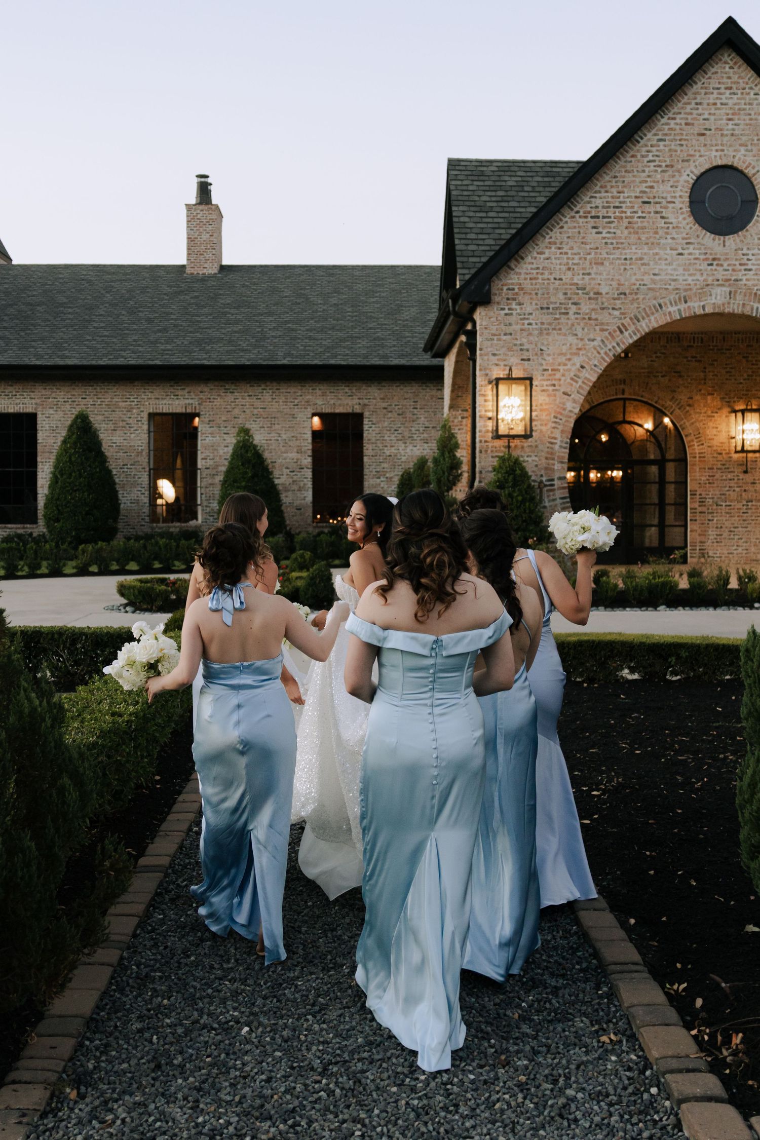 Bride and bridesmaids in light blue satin gowns walk toward an elegant brick venue at dusk.