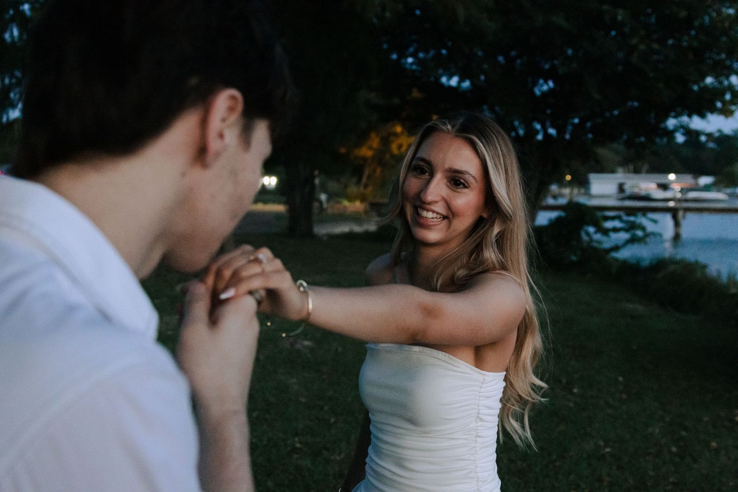 Smiling blonde bride in white strapless dress holds groom's hand in evening garden setting.