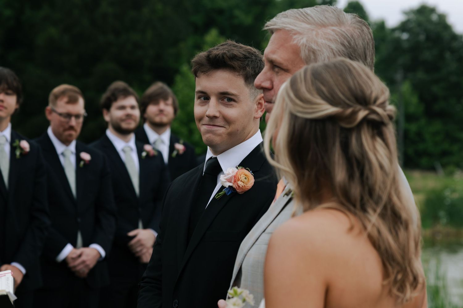 Young groomsman in black tuxedo smiling during outdoor wedding ceremony with groomsmen lined up behind.
