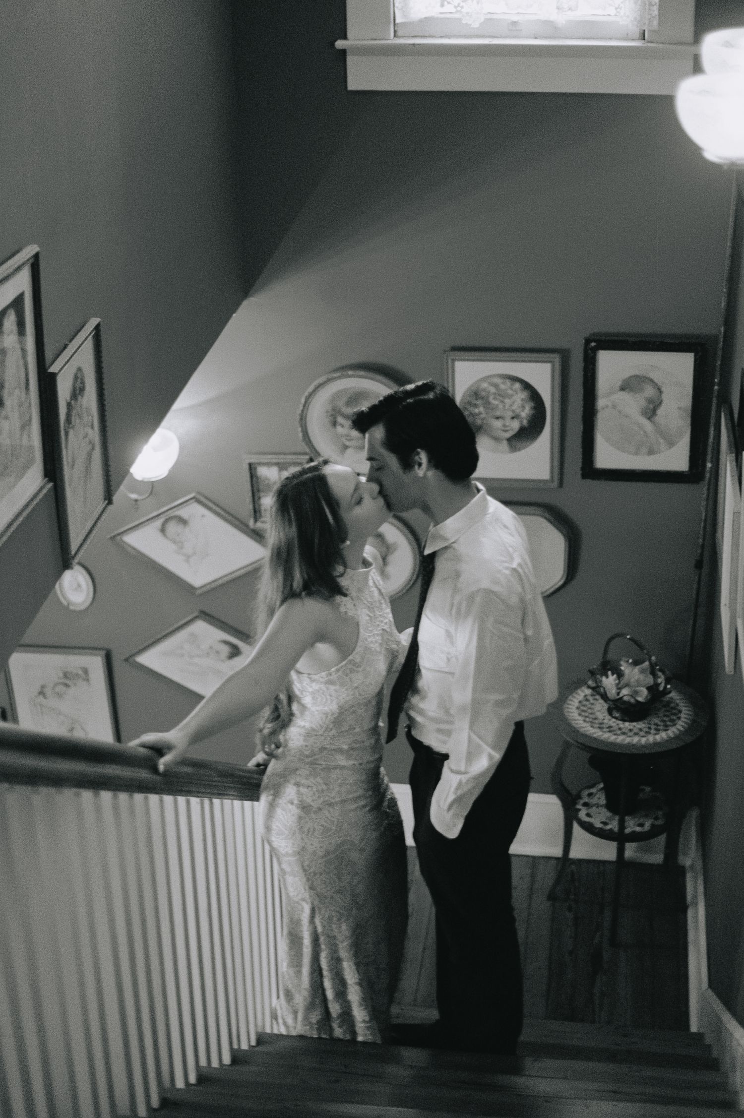 Black and white photo of bride and groom kissing on a staircase surrounded by framed wall art.