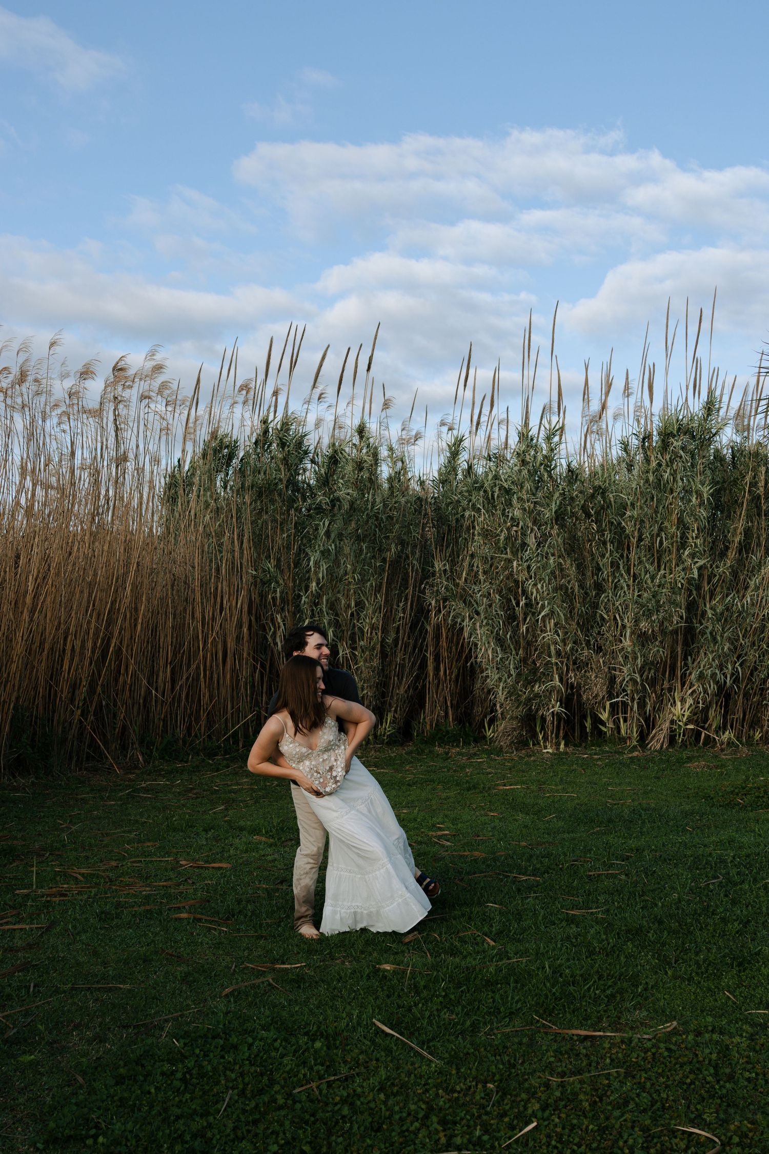 Woman in white dress spinning playfully in a grassy field with tall dry reeds under a blue sky.