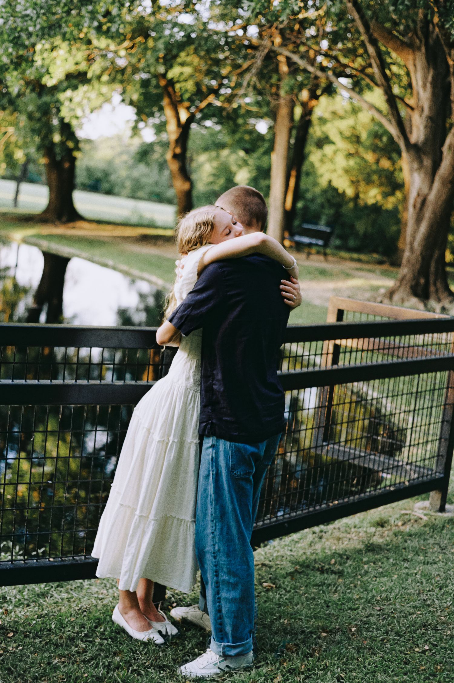 Couple embracing in a romantic outdoor setting near a pond, surrounded by lush green trees.