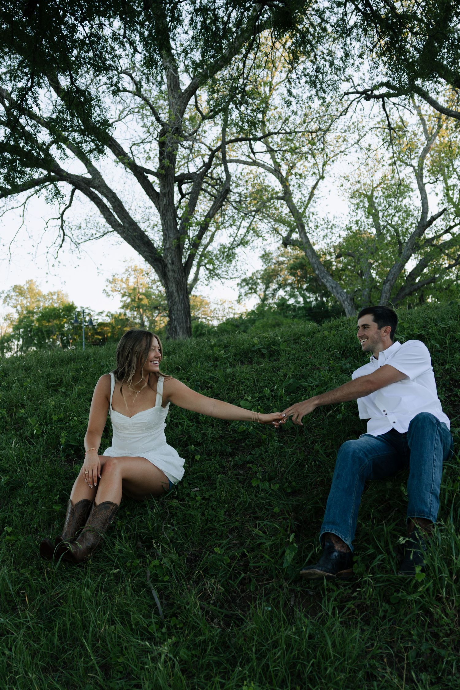 Couple holding hands while sitting on a grassy hillside during an outdoor engagement photo session.