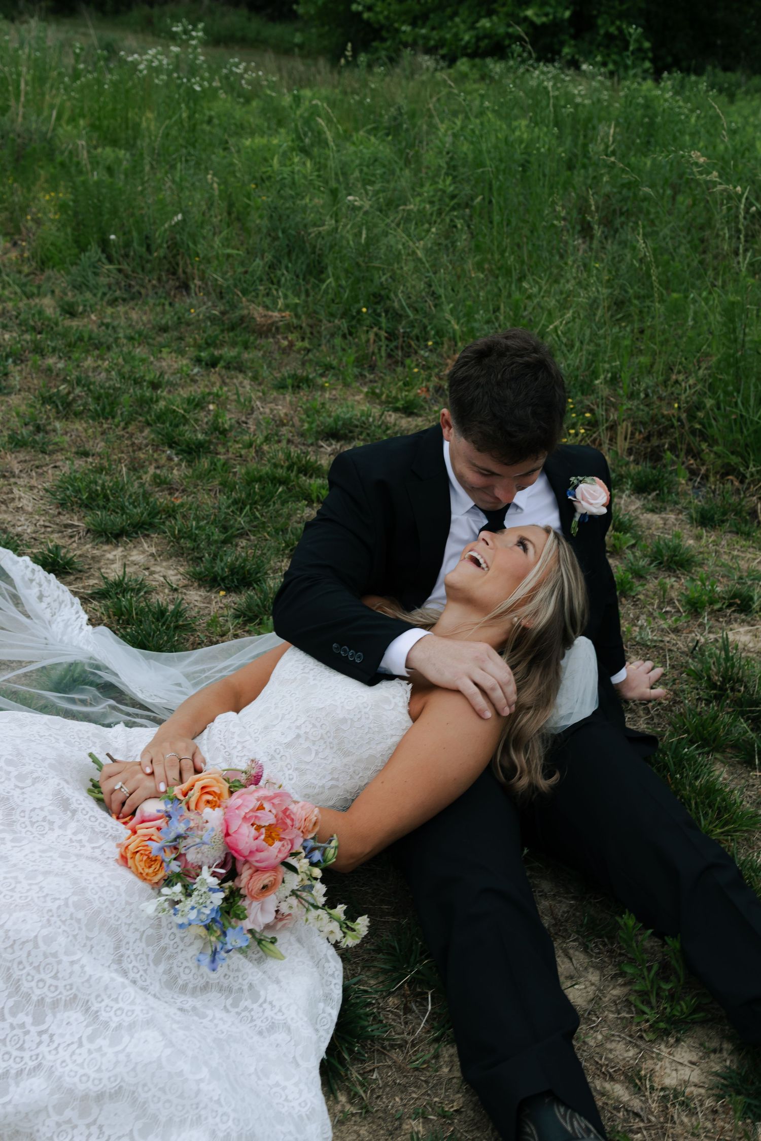 Bride and groom sit in grass, bride leaning back holding colorful bouquet while groom embraces her tenderly.