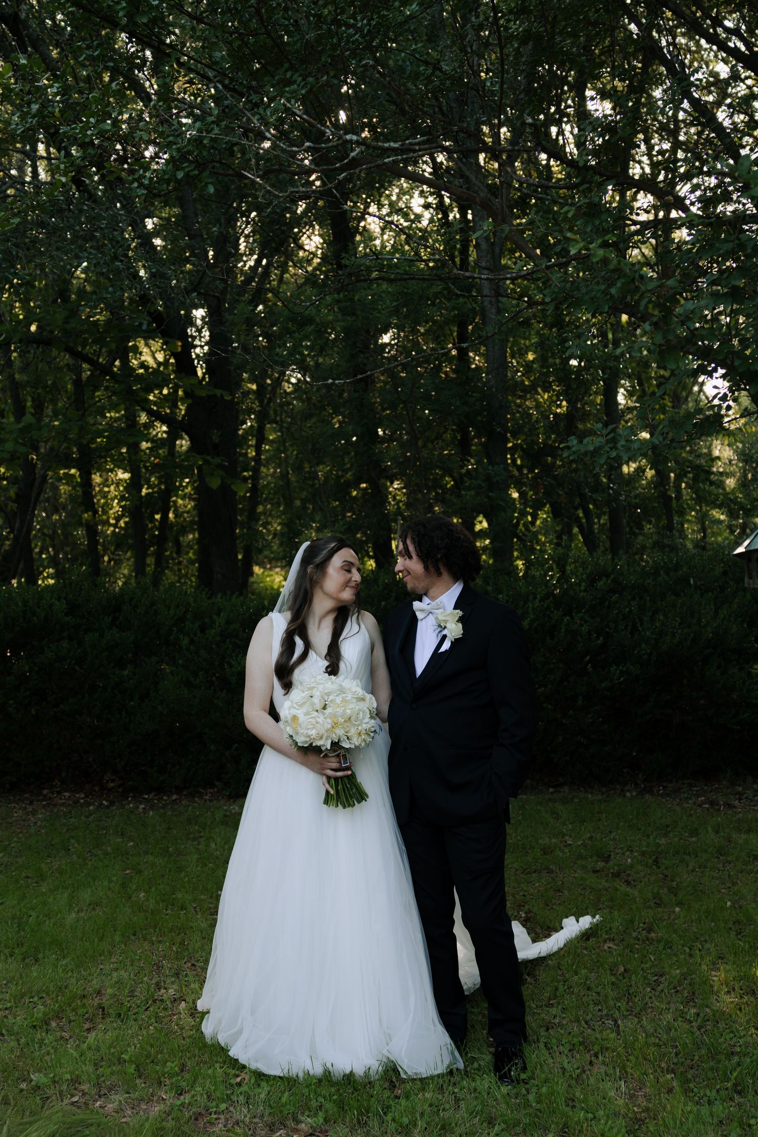 Bride in white tulle gown and groom in black suit stand face to face in a lush green wooded outdoor setting.