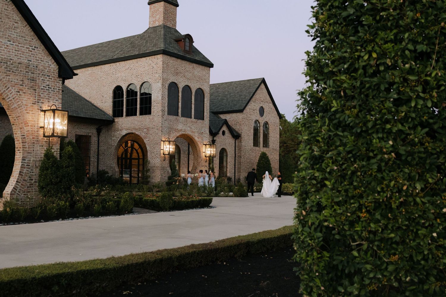 Stunning brick mansion wedding venue at dusk with warm lighting, manicured hedges, and bride in white gown.