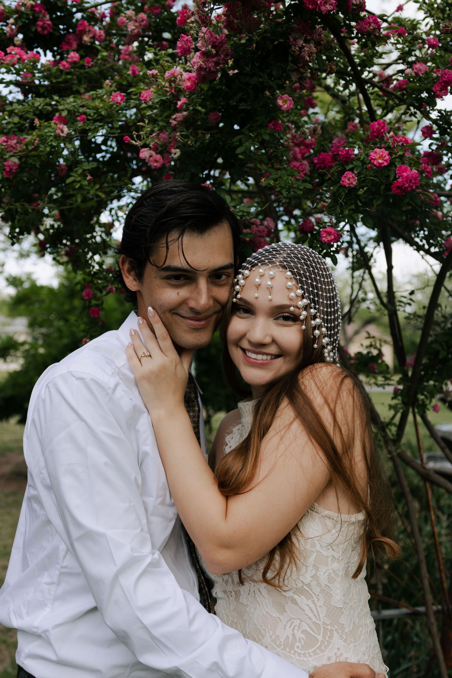 Smiling bride in pearl veil embraces groom under pink flowering roses outdoors.