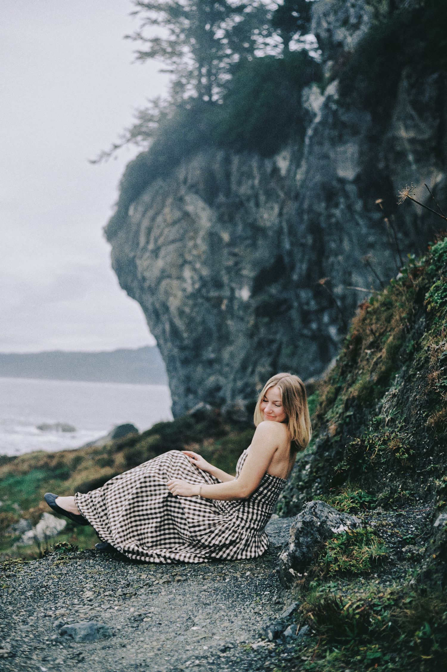 Woman in a gingham dress sitting peacefully against a mossy cliff with ocean views in the background.