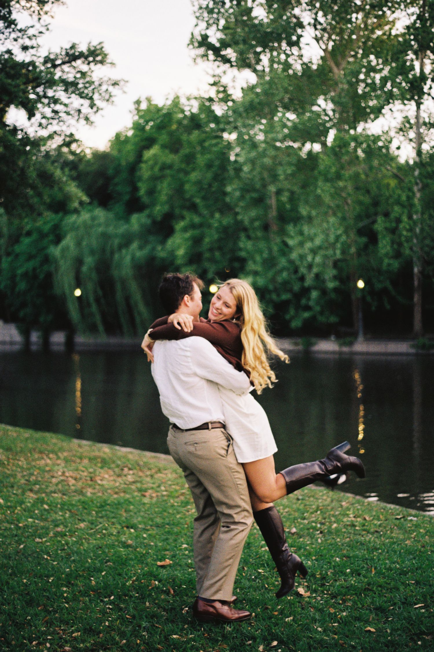 Couple embracing by a park lake, man lifting woman in brown boots and white dress among green trees.