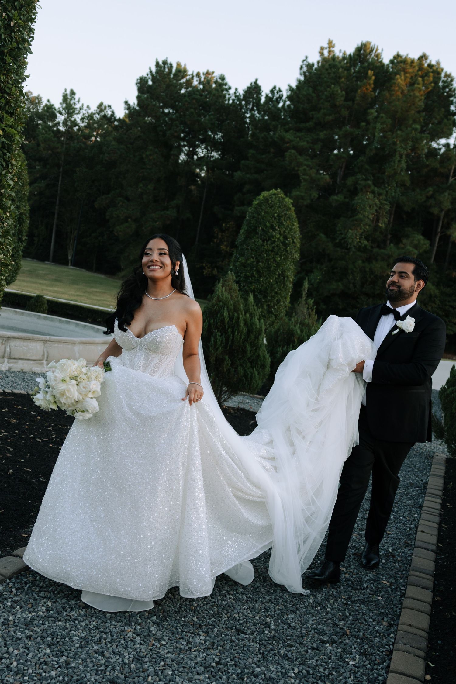Bride in sparkling ball gown holding white bouquet while groom in tuxedo carries her train outdoors.