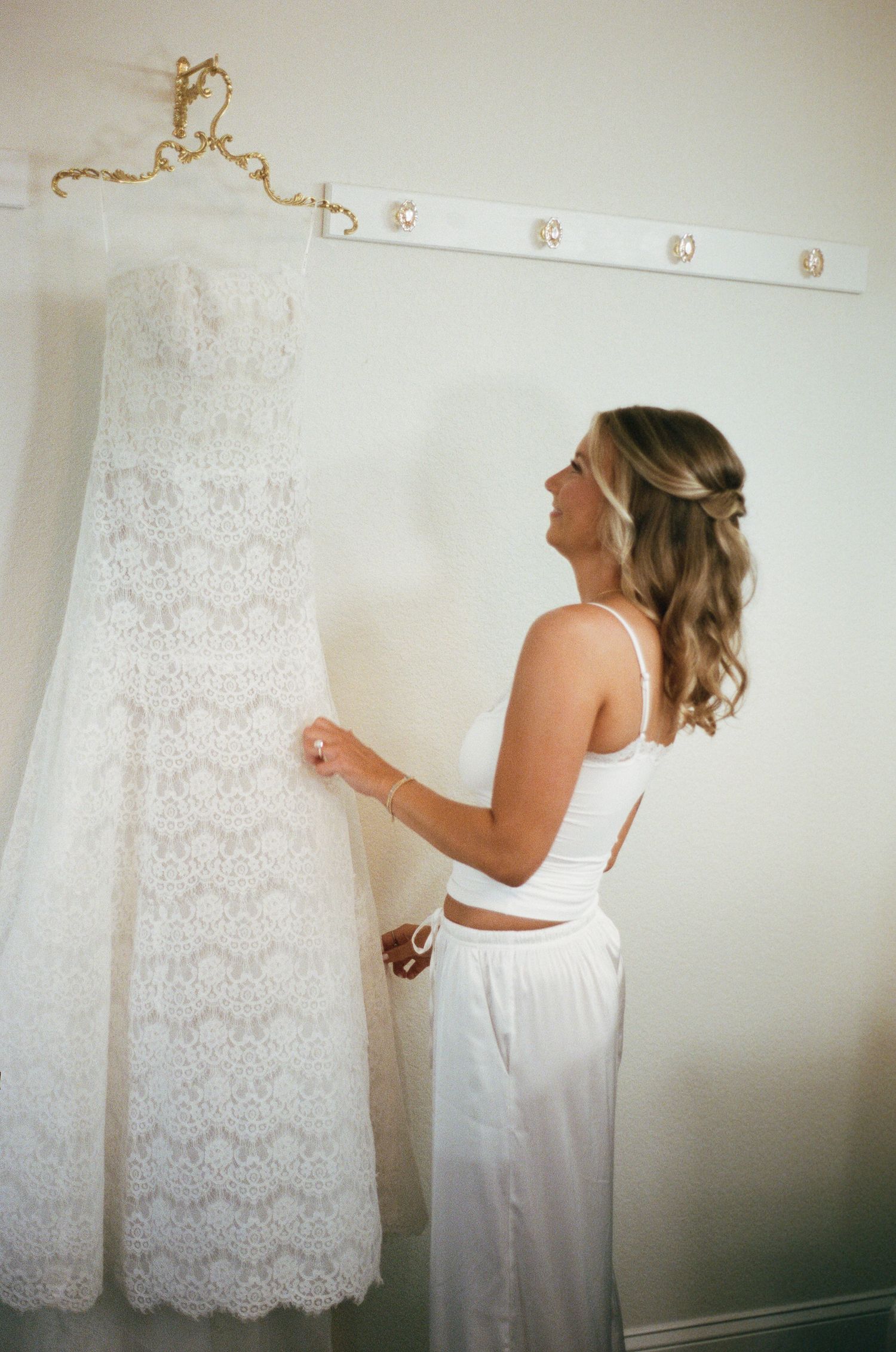 Bride admiring her lace wedding gown hanging on a wall hook while dressed in white separates.