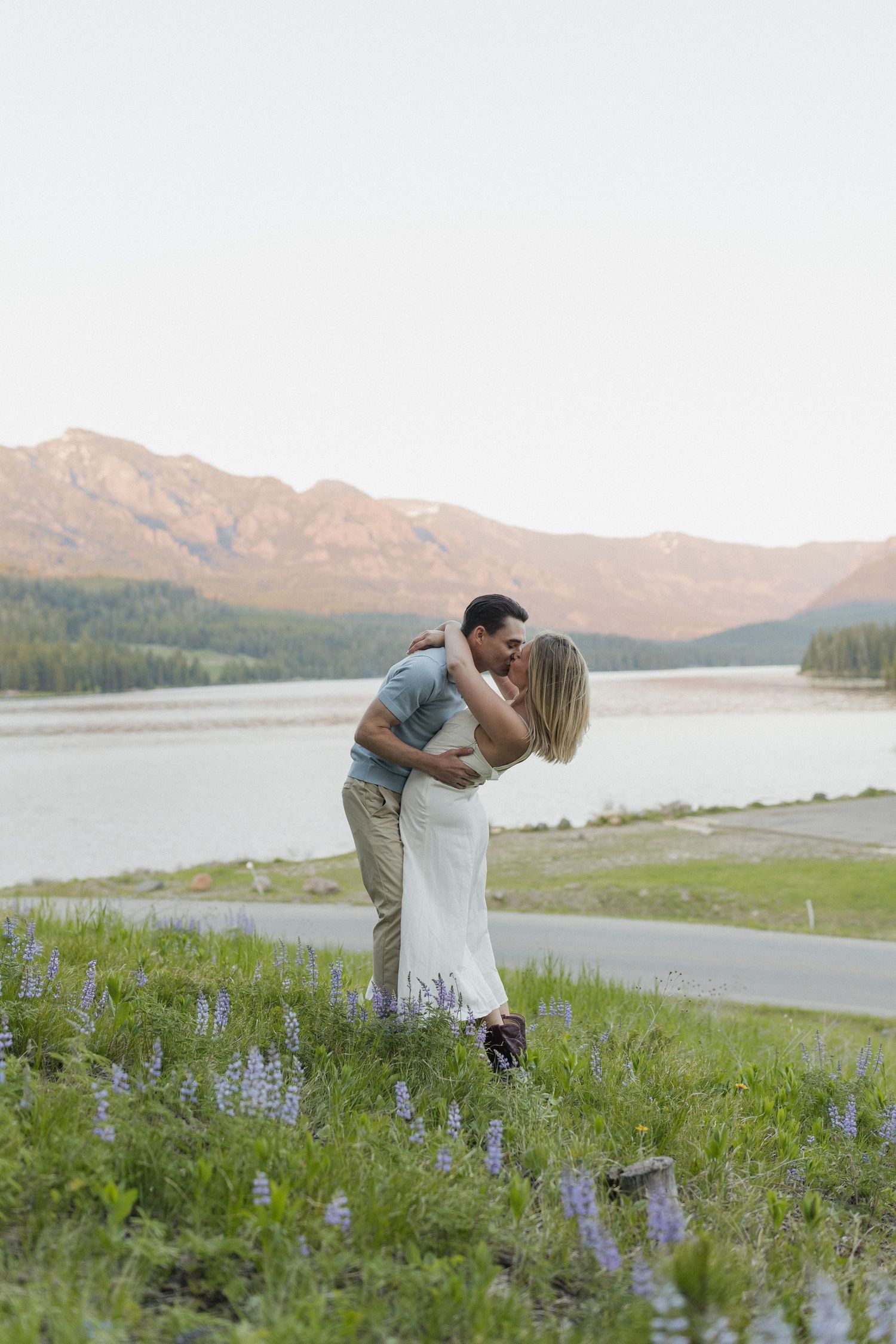 A romantic embrace on a grassy hillside with purple wildflowers and a mountain lake view.