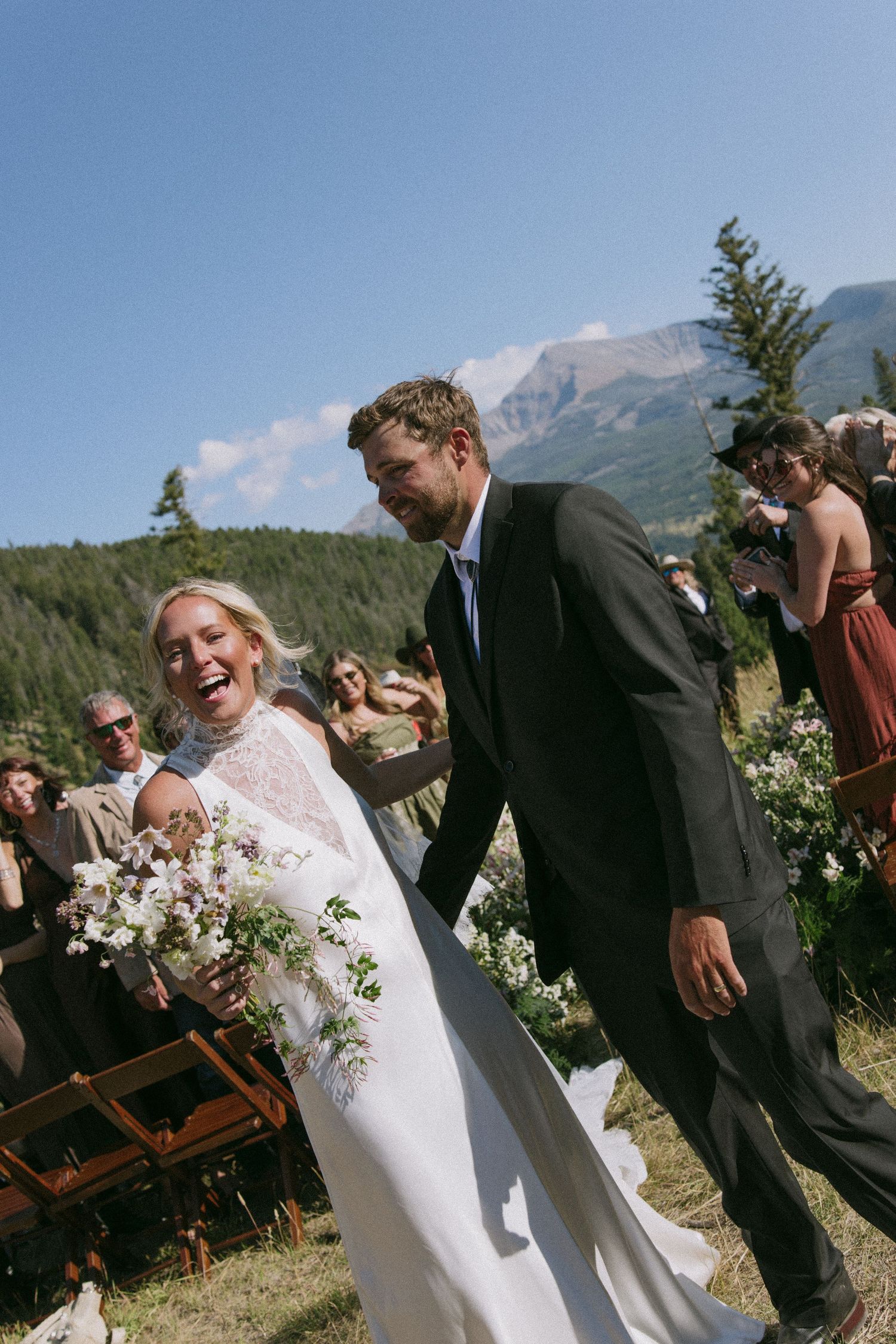 A joyful outdoor wedding ceremony takes place against a scenic mountain backdrop with greenery and blue skies.