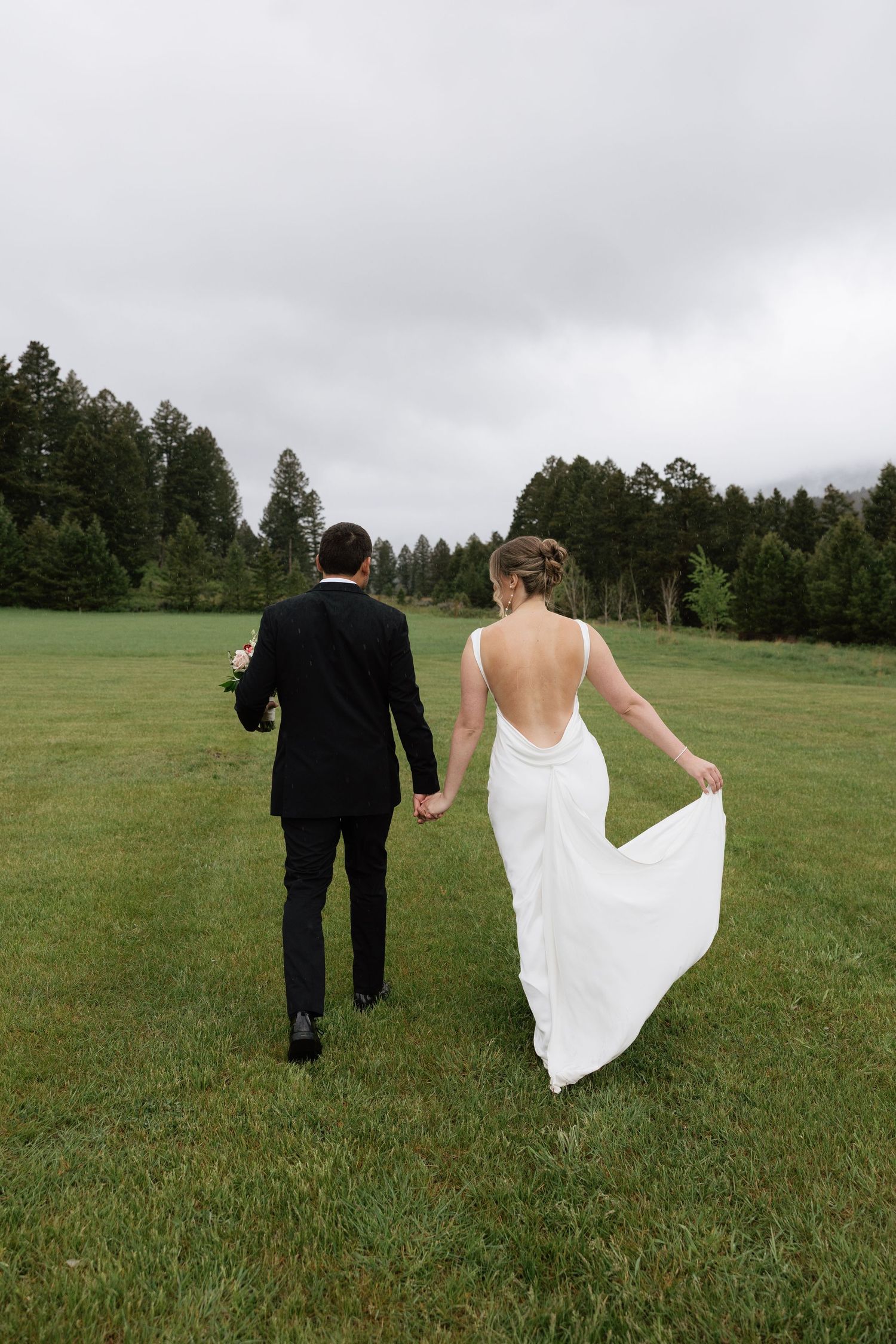 Bride and groom walking hand in hand across green meadow, bride in backless white gown, groom in black suit, pine trees behind.