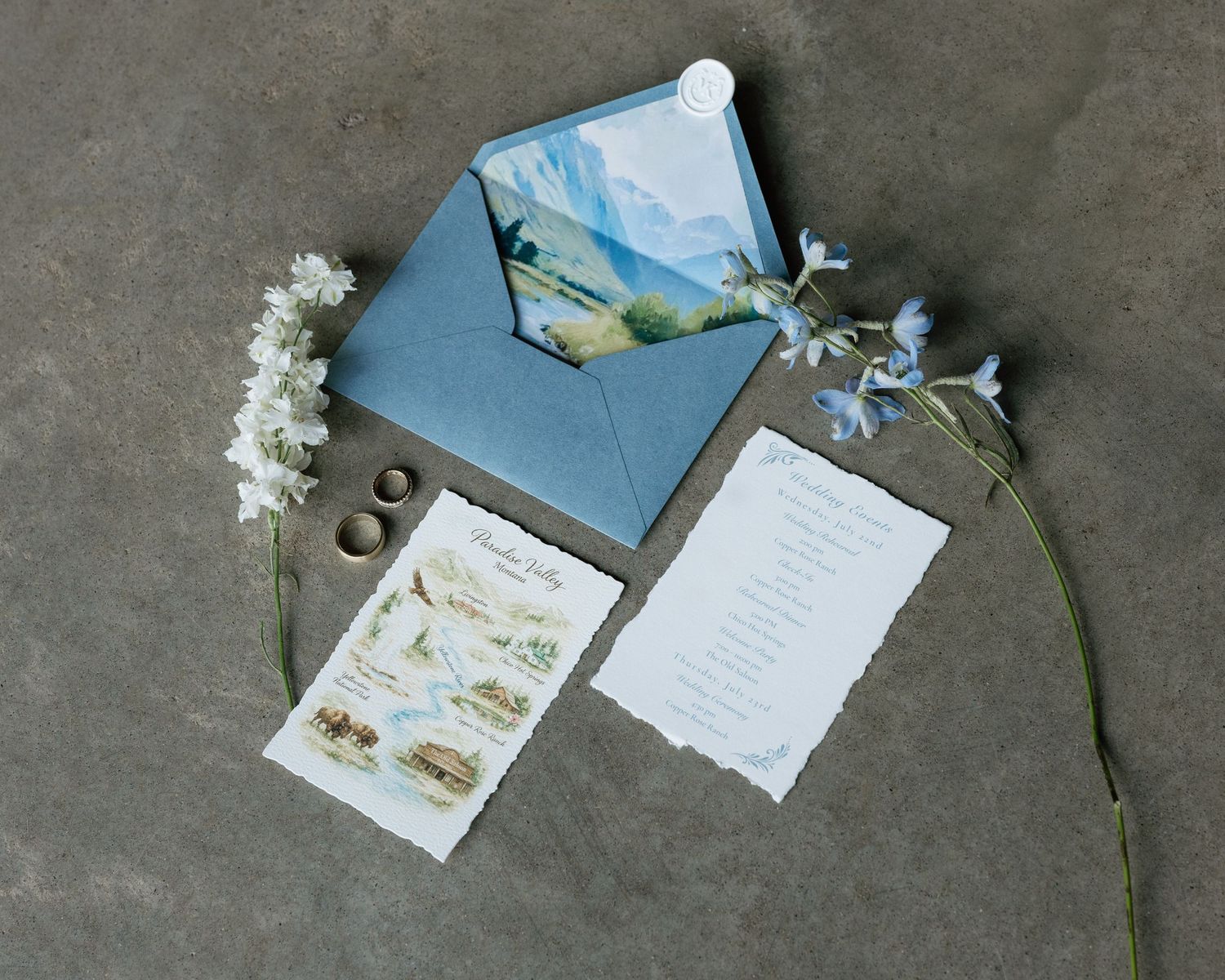 Blue envelope wedding stationery suite with watercolor mountain liner, floral accents, rings, and deckled edge paper on gray background.