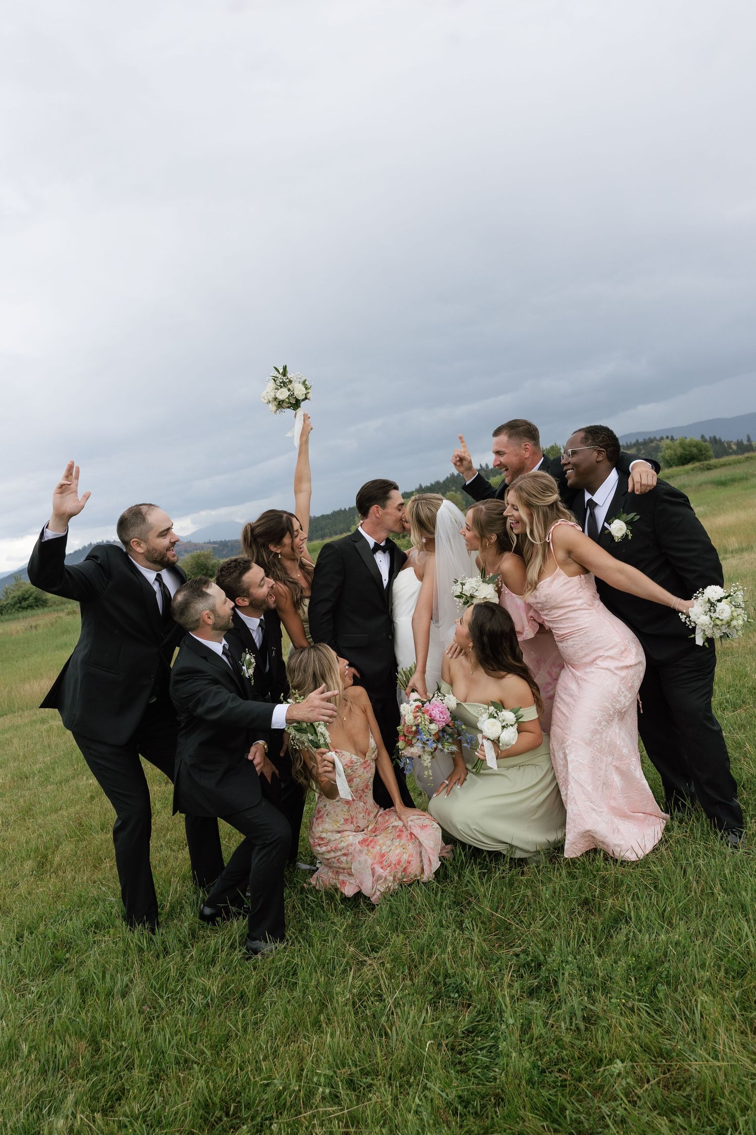 Wedding party celebrating outdoors on green grass, bridesmaids in pink and sage gowns, groomsmen in black tuxedos, bride tossing bouquet.