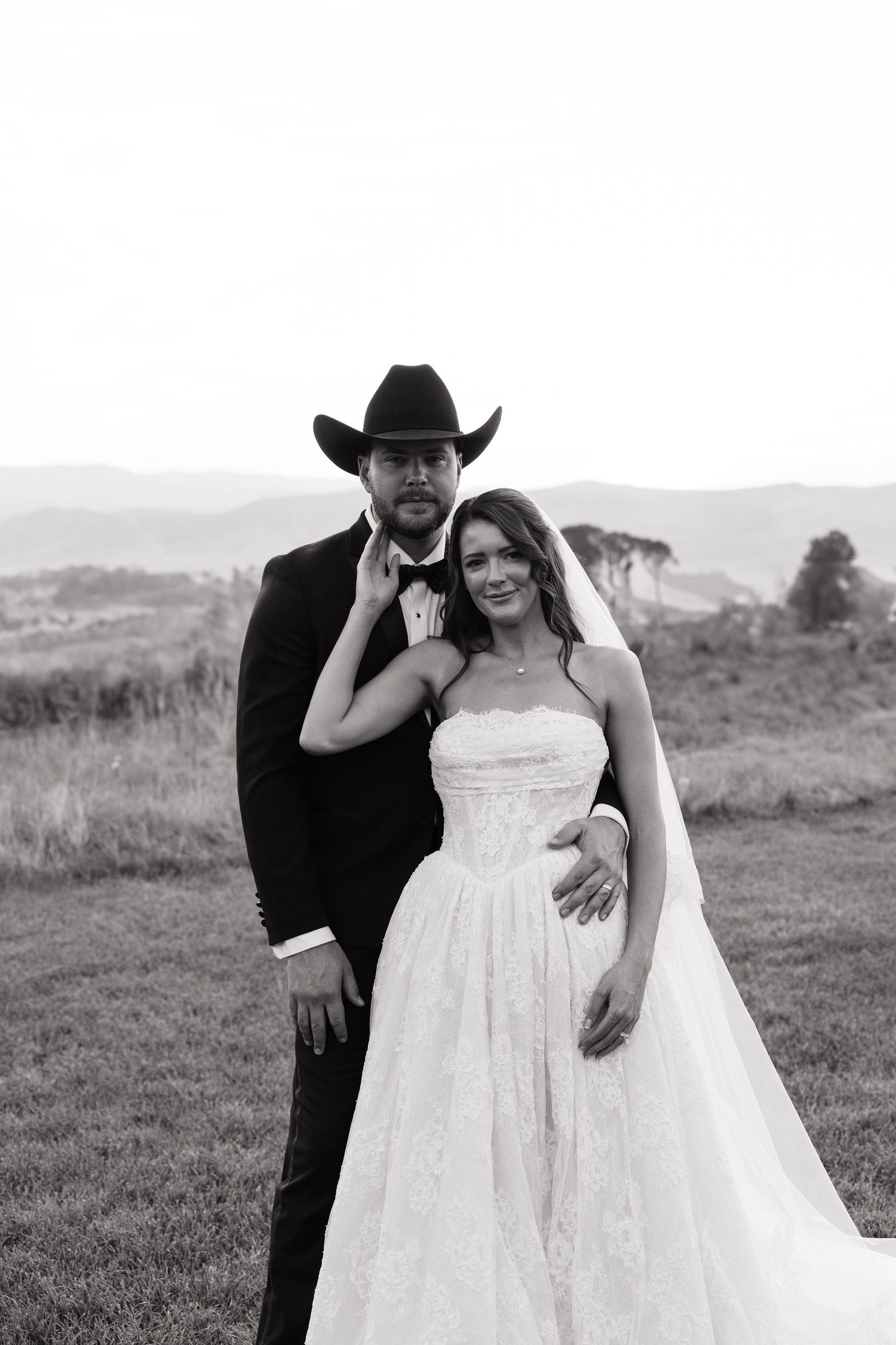 Black and white wedding photo of bride in strapless gown and groom in tuxedo with cowboy hat, posing outdoors.