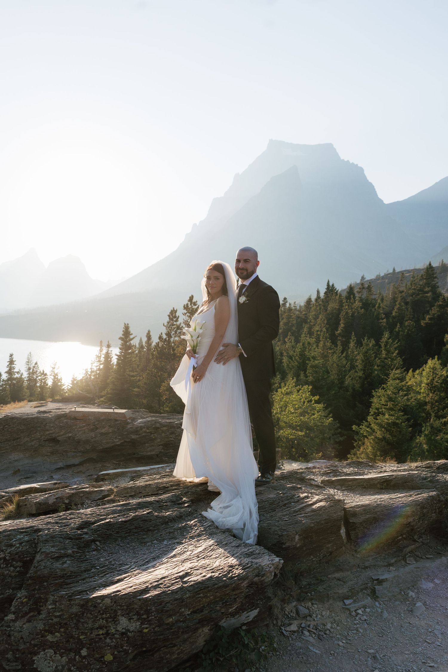 Bride and groom posing on rocky ledge with mountain lake and evergreen forest backdrop at golden hour.