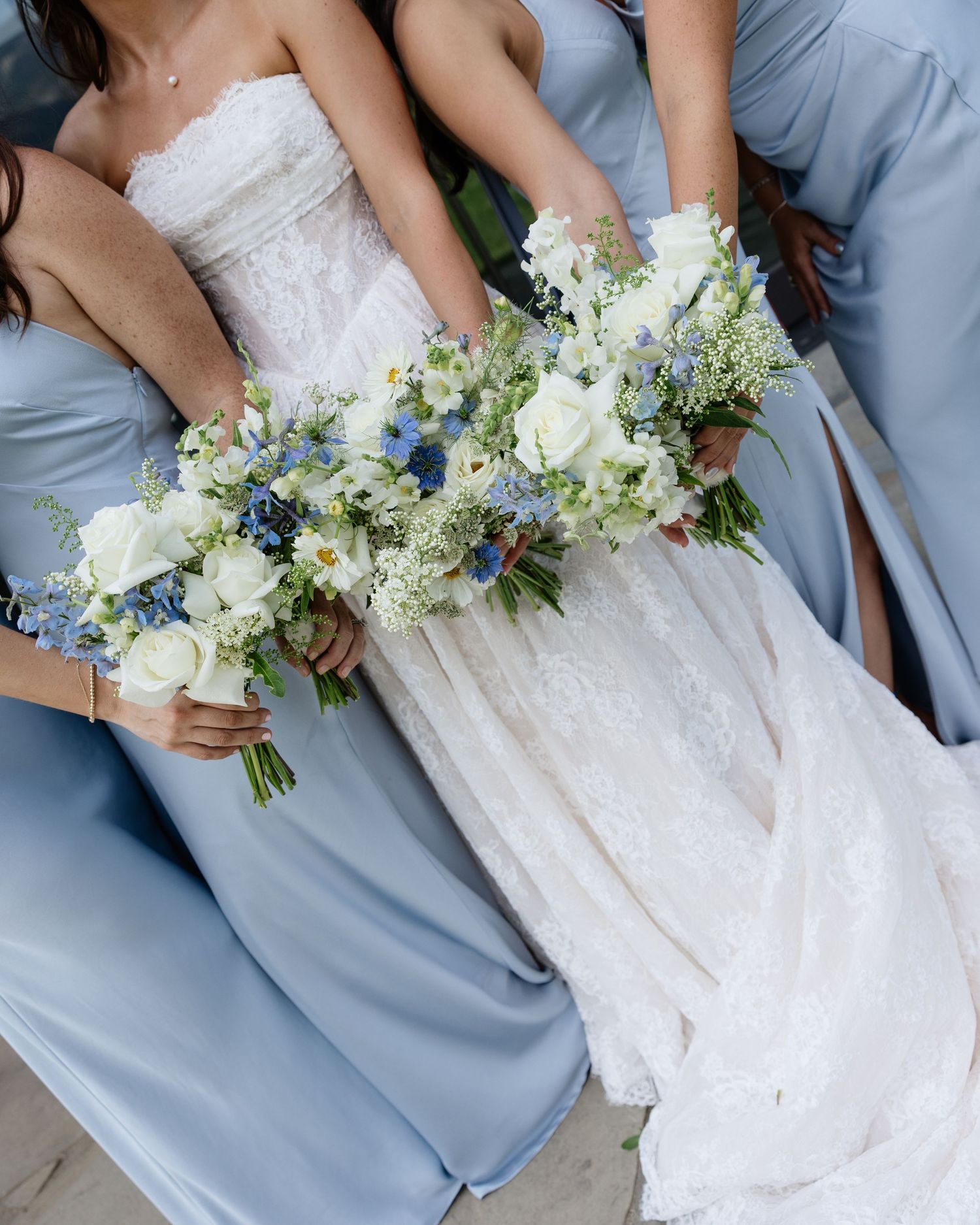 Bride in white lace gown with bridesmaids in blue dresses holding white and blue floral bouquets.