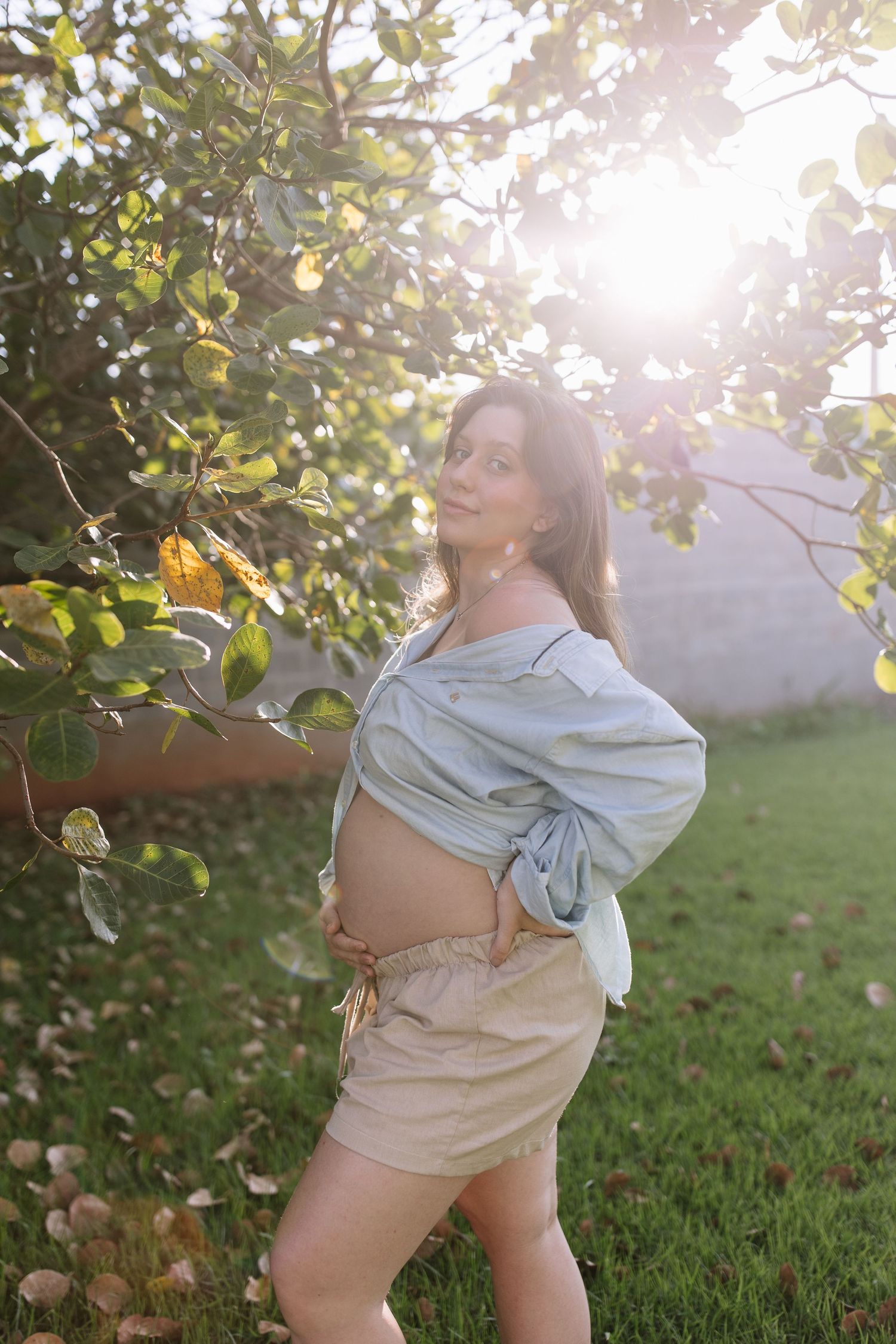 Sunlit maternity portrait in a garden with blooming trees and soft natural lighting.