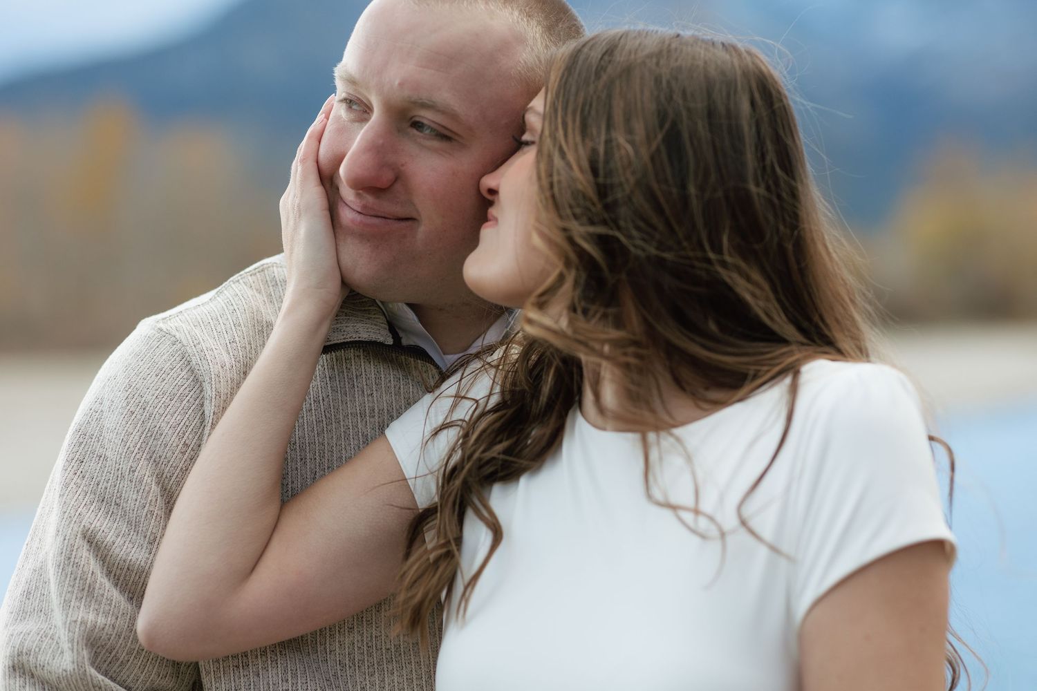 A woman tenderly kisses a smiling man on the cheek outdoors with mountains in the background.