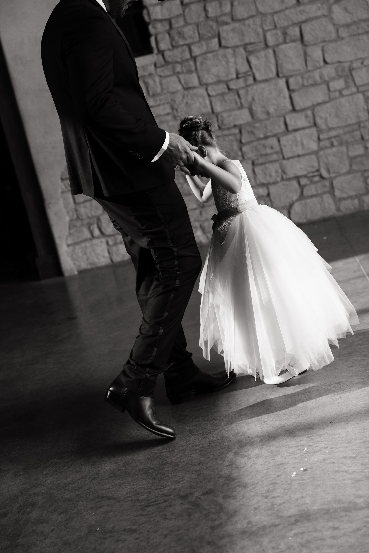 Black and white photo of a man in a suit dancing with a little girl in a white dress at a wedding reception.