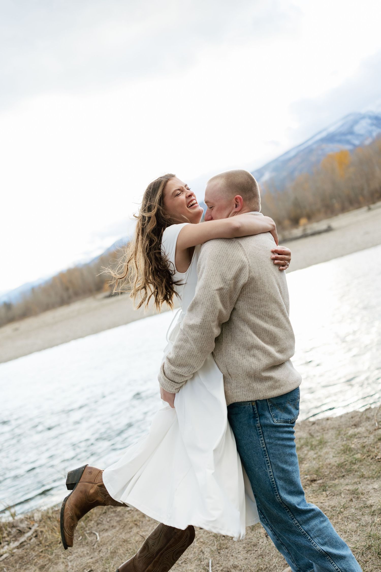 Couple laughing and embracing near a mountain lake, woman in white dress and brown boots, man in beige sweater and jeans.