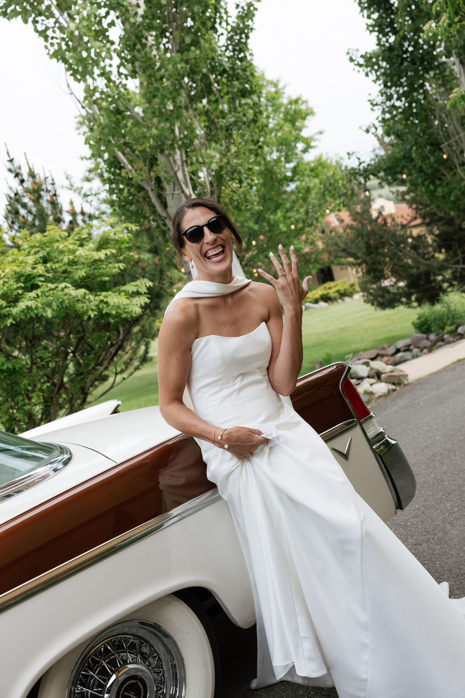Smiling bride in white strapless gown and sunglasses showing ring while leaning on classic vintage white car outdoors.