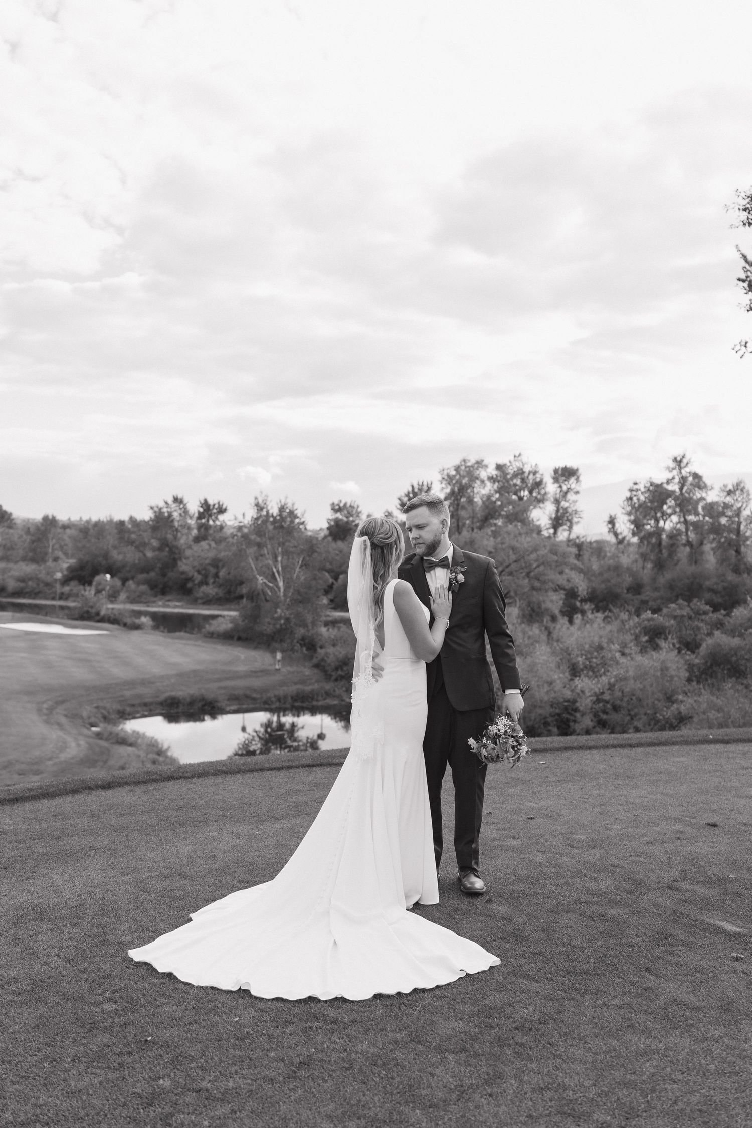 Black and white photo of bride and groom standing on golf course, bride in flowing gown with veil, groom in suit.