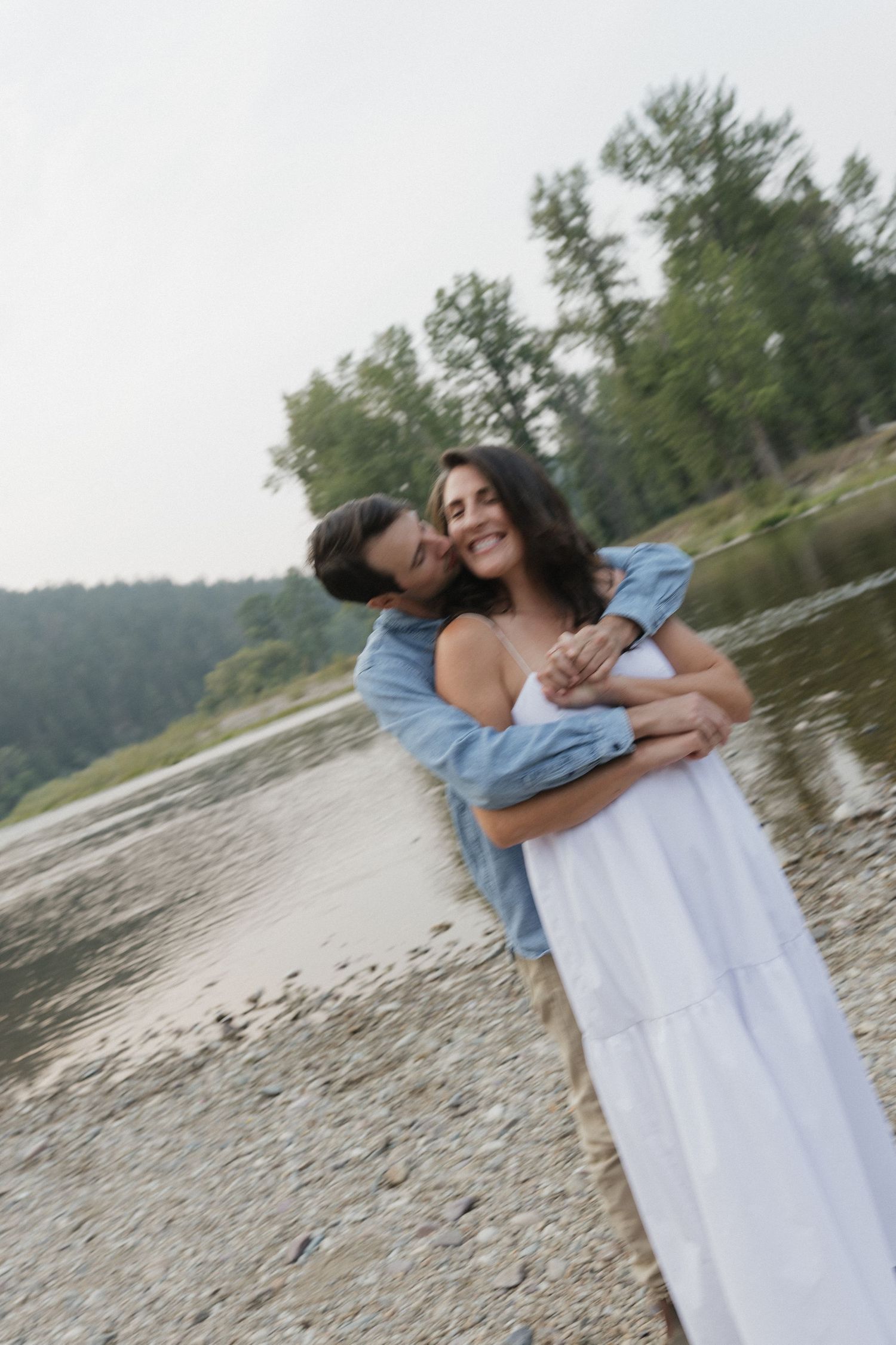 A couple embracing by a scenic lake with trees in the background.