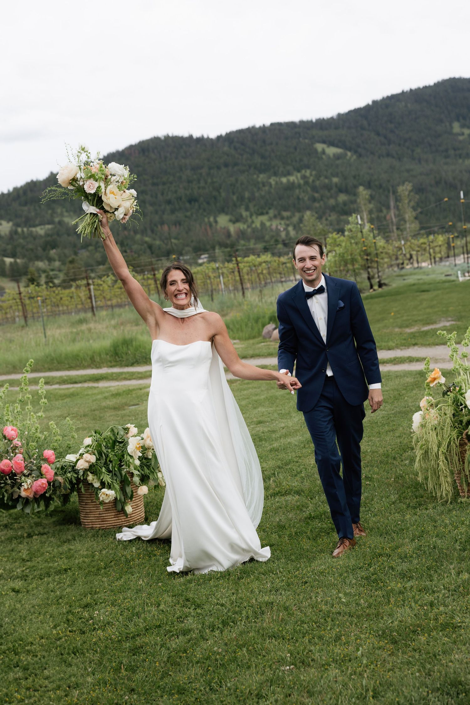 Joyful bride raises bouquet while groom in navy suit holds her hand at outdoor mountain vineyard wedding ceremony.