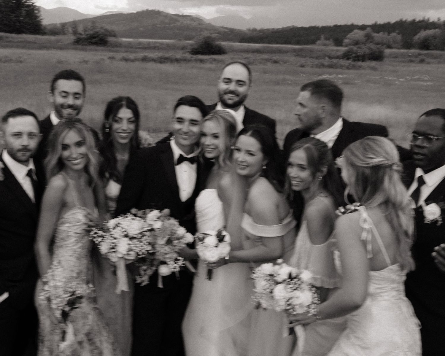 Black and white wedding party photo outdoors with mountains, bride in white gown, groom in tuxedo, surrounded by bridesmaids and groomsmen.