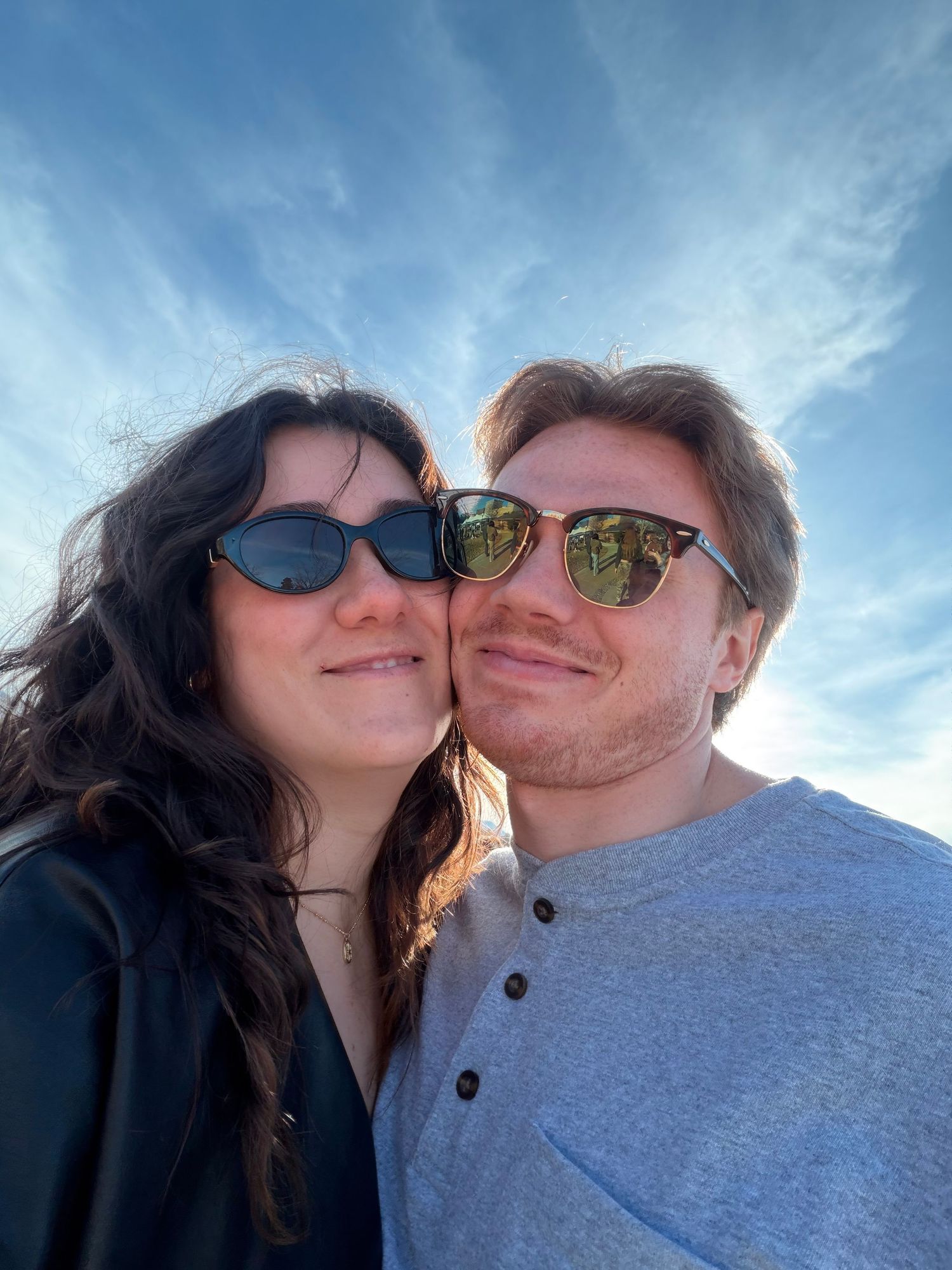 A couple wearing sunglasses takes a series of selfies together against a bright blue sky background.