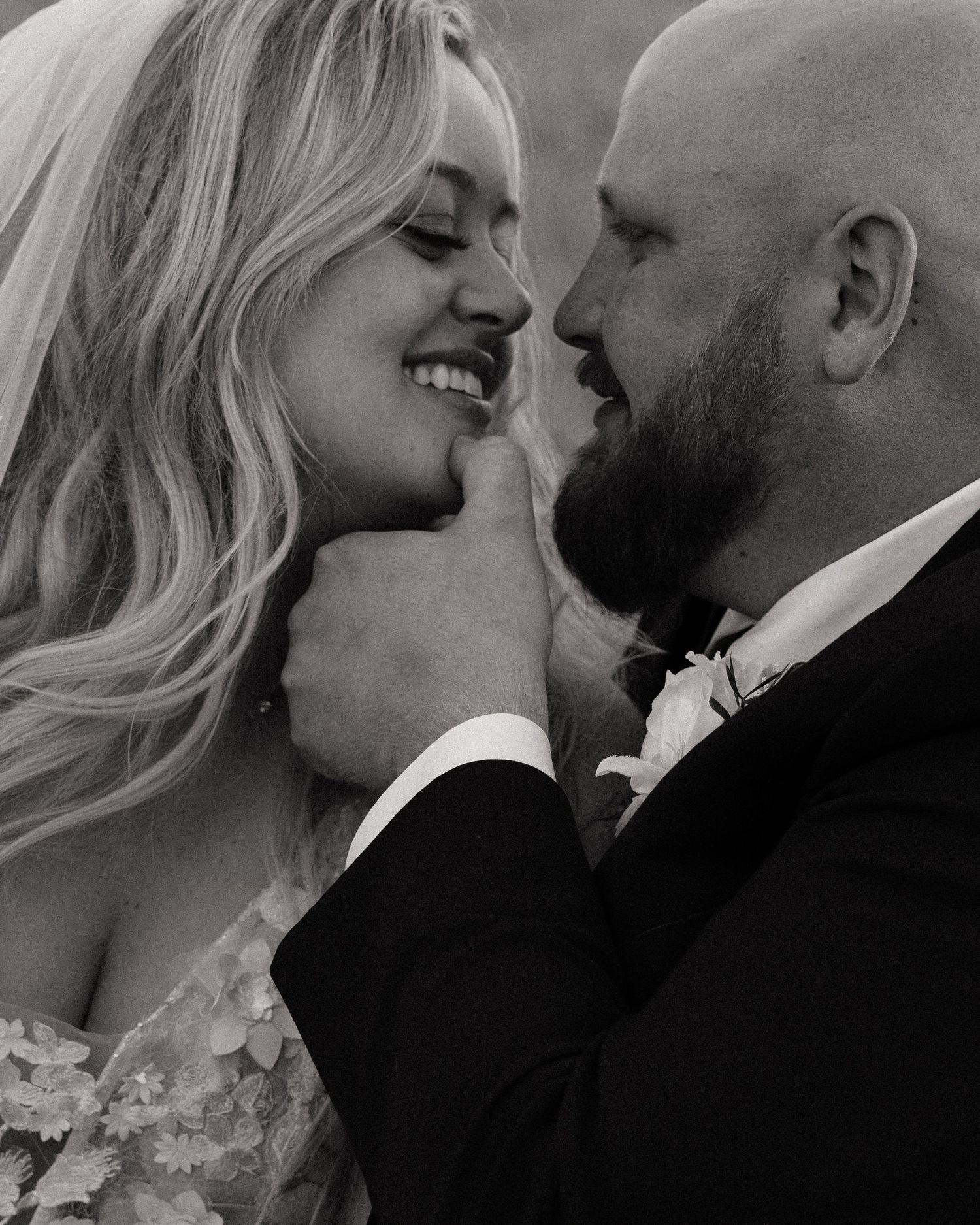 Black and white wedding photo of smiling bride and bearded groom in tuxedo sharing an intimate moment.
