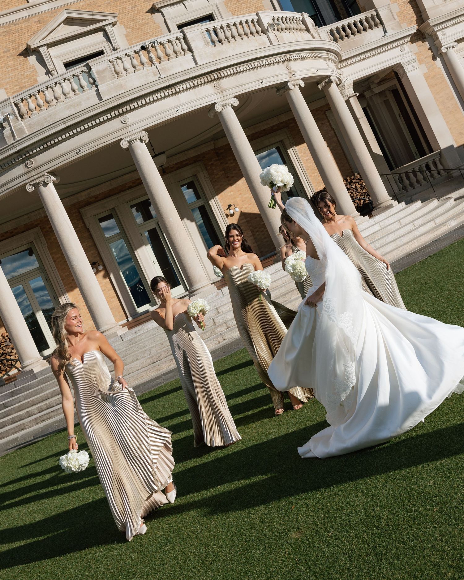 Wedding party celebrates on manicured lawn in front of grand columned building entrance.