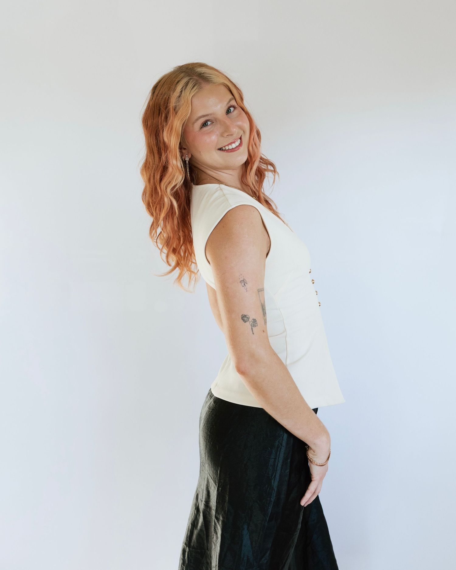 Woman with red wavy hair smiling, wearing cream top and dark skirt, showing arm tattoos, posing sideways.