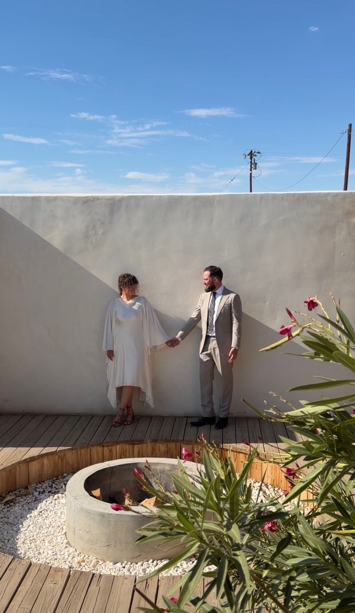 Couple in wedding attire holding hands on a wooden rooftop terrace with white walls, tropical plants, and blue sky.